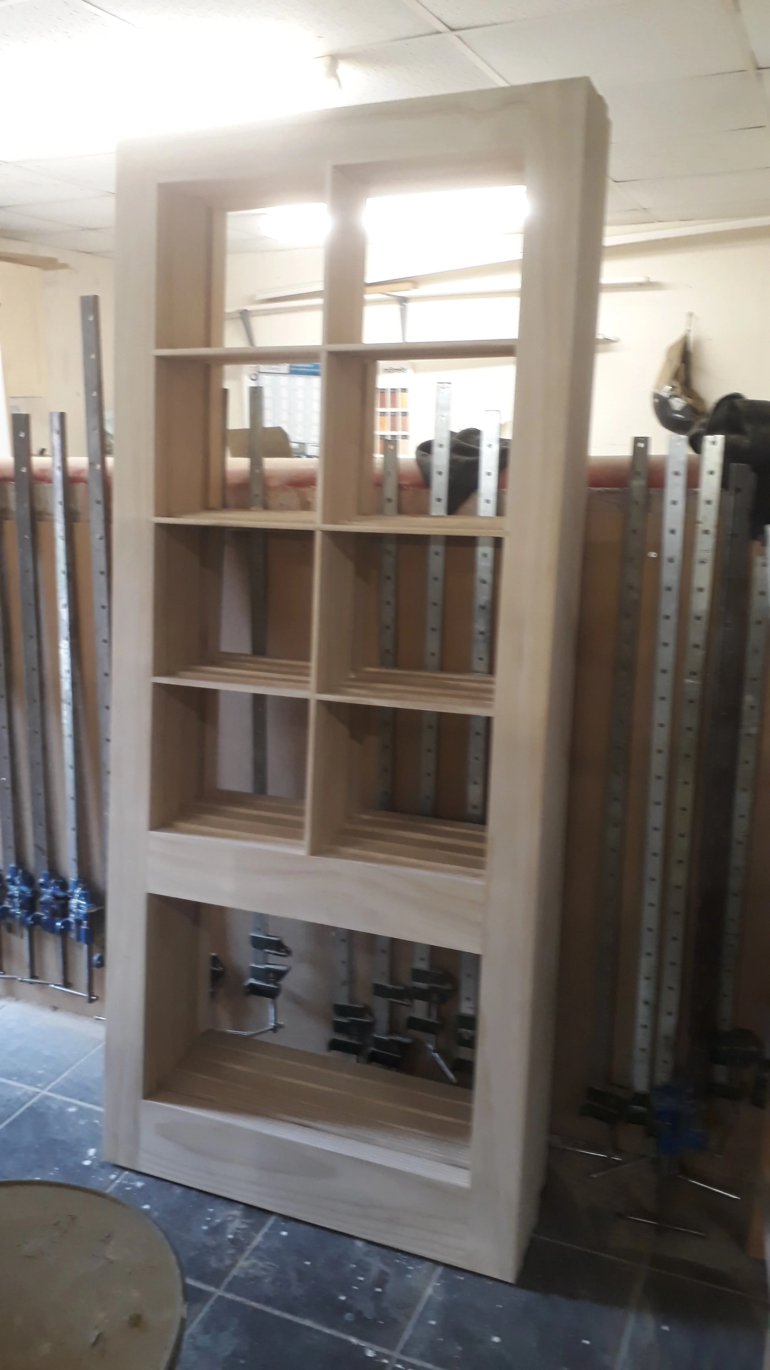 A wooden bookshelf with multiple empty compartments standing in a workshop, surrounded by clamps and woodworking tools.