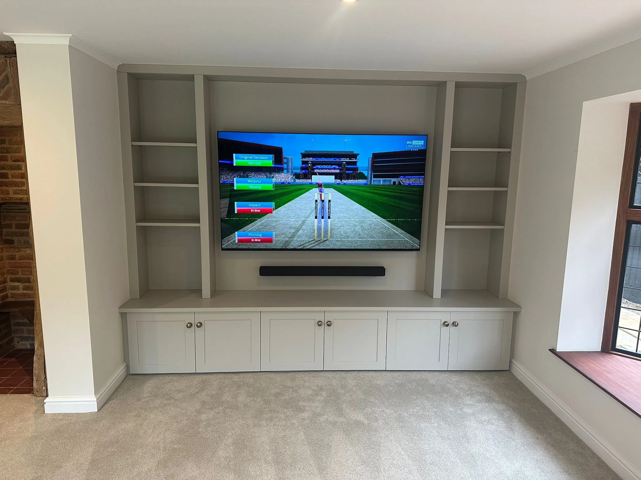 Living room entertainment center with large flat-screen TV displaying a cricket game, surrounded by empty white shelves and cabinets, with a window on the right side.