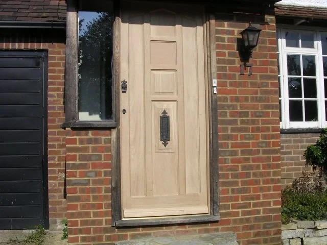 A beige front door with decorative paneling and a mail slot, set in a brick wall next to a window, with a black lantern-style light fixture beside it.