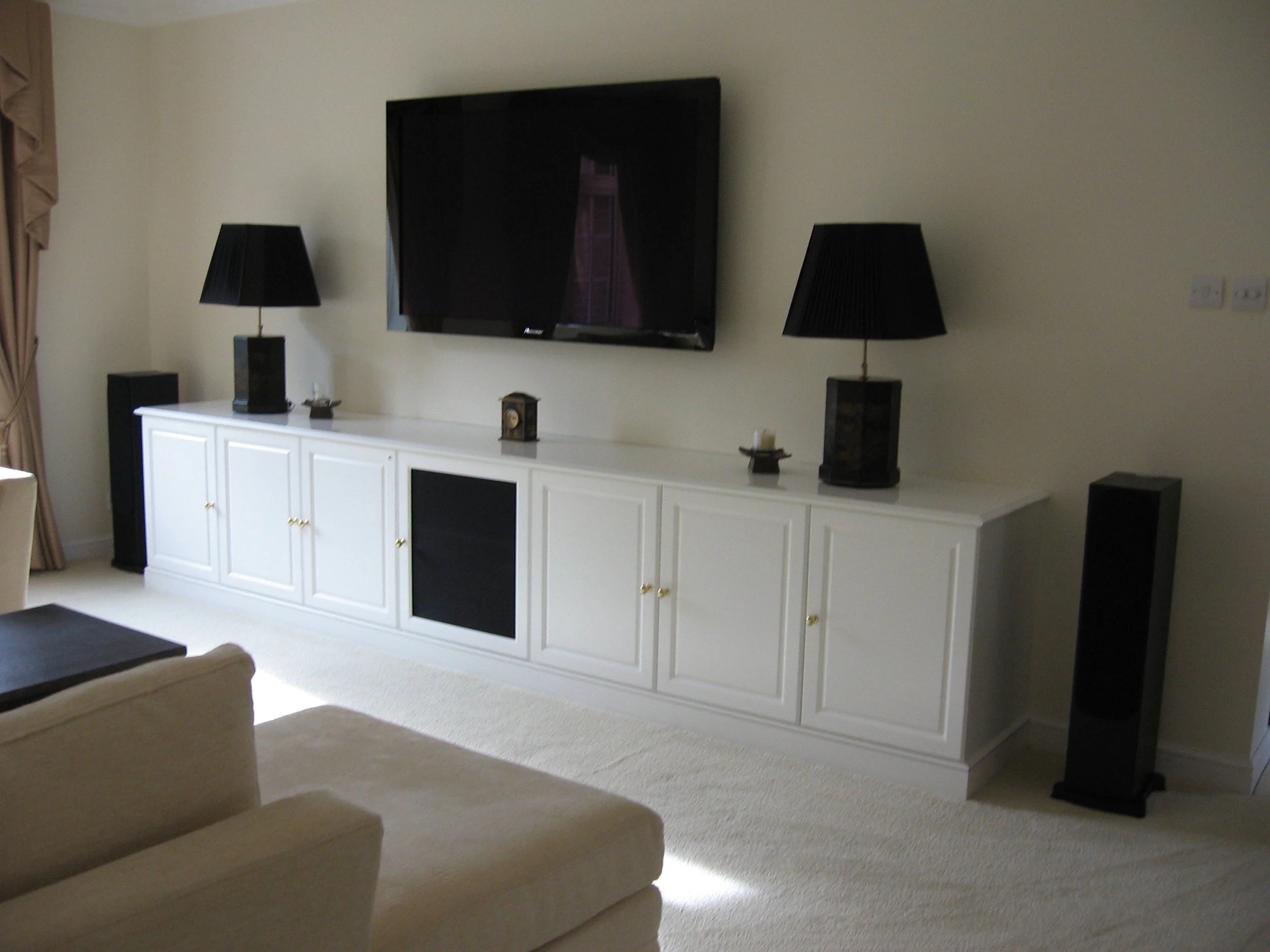 Living room with a wall-mounted flat-screen TV, white cabinet with gold handles, black table lamps, and speakers.