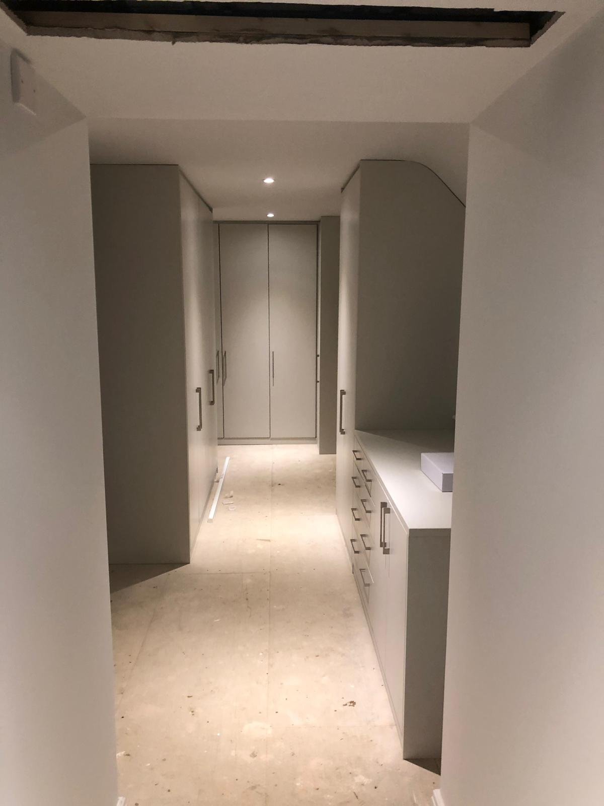 A corridor with white built-in cabinets on both sides, a beige tiled floor, and ceiling recessed lighting, leading to closed doors at the end.