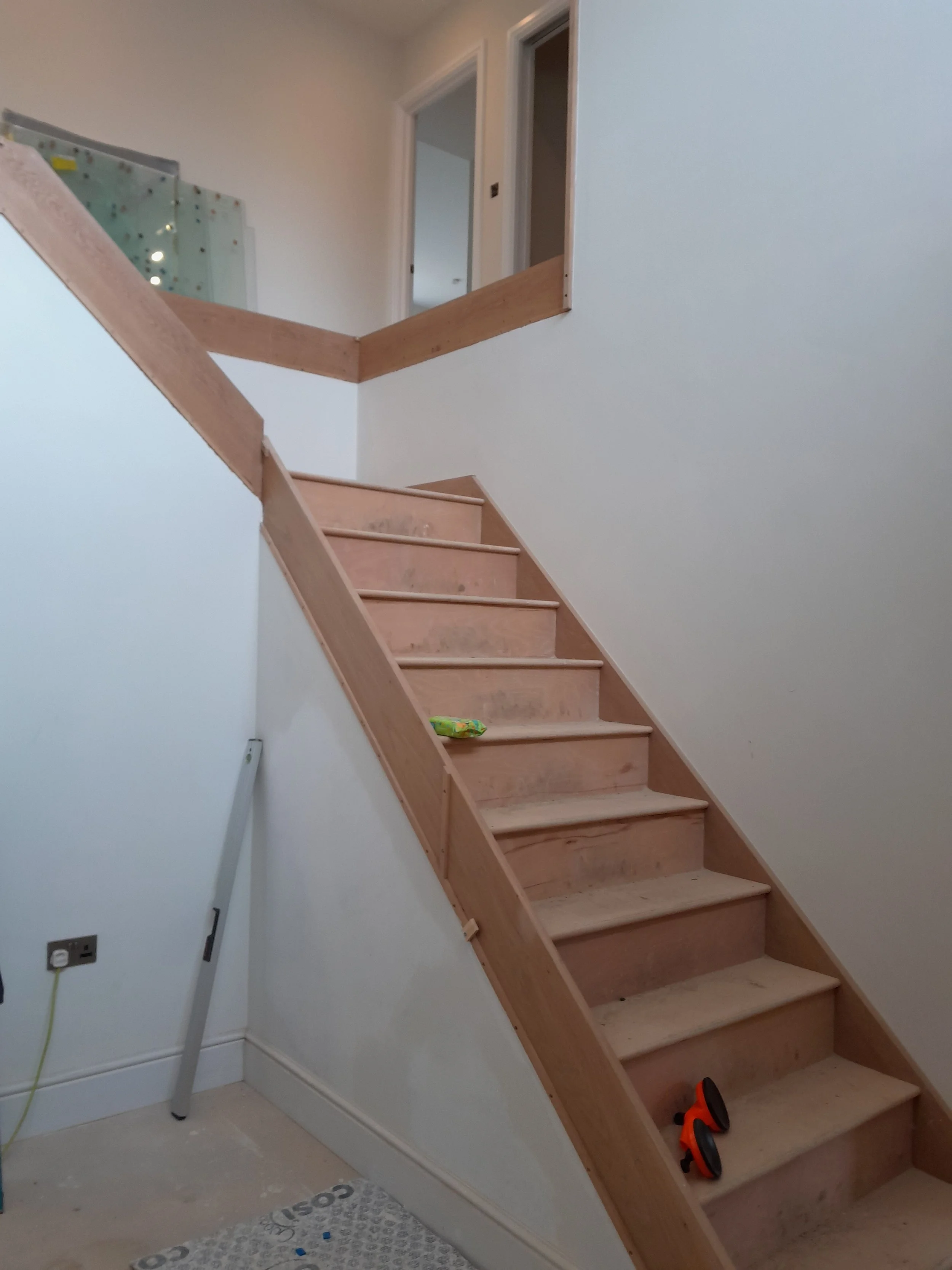 Unfinished wooden staircase with a pair of orange and black shoes on one step, a green snack wrapper, and construction tools, leading upstairs in a house under renovation.