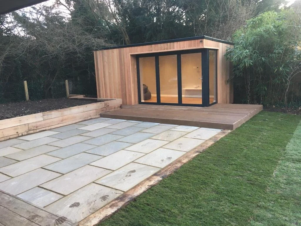 A small wooden building with large sliding glass doors, set on a wooden deck, surrounded by grass and trees.
