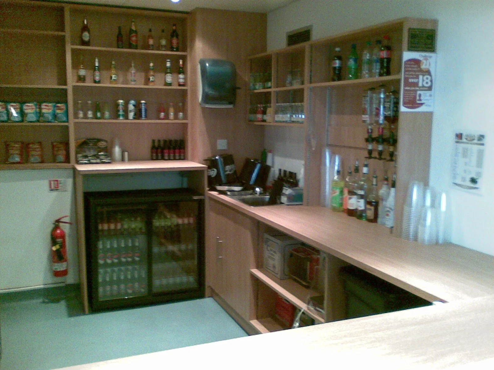 A bar area with wooden shelves stocked with bottles of alcohol and snacks, a small refrigerator, and a bar counter with cups and bar supplies.