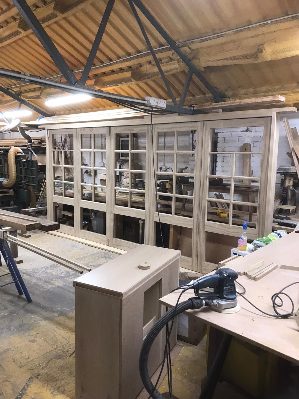 Woodworking workshop with a partially assembled wooden window frame and various tools, including a sander, on workbenches.