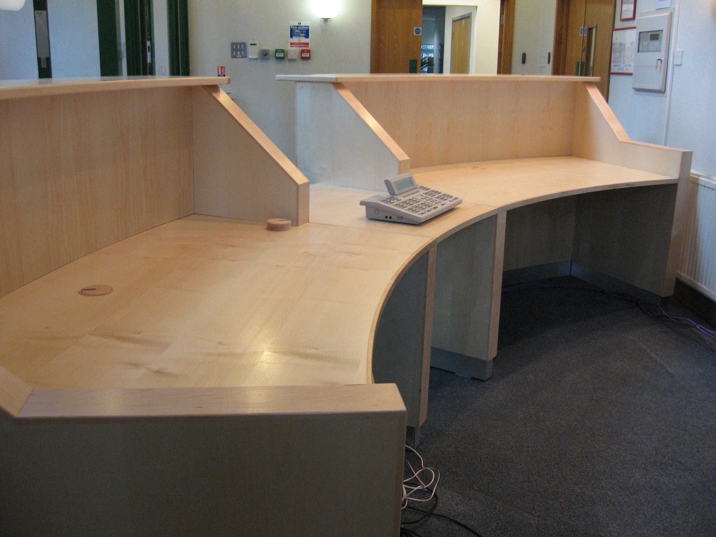 Empty wooden reception desk with a telephone and some electrical outlets, in a well-lit office space.