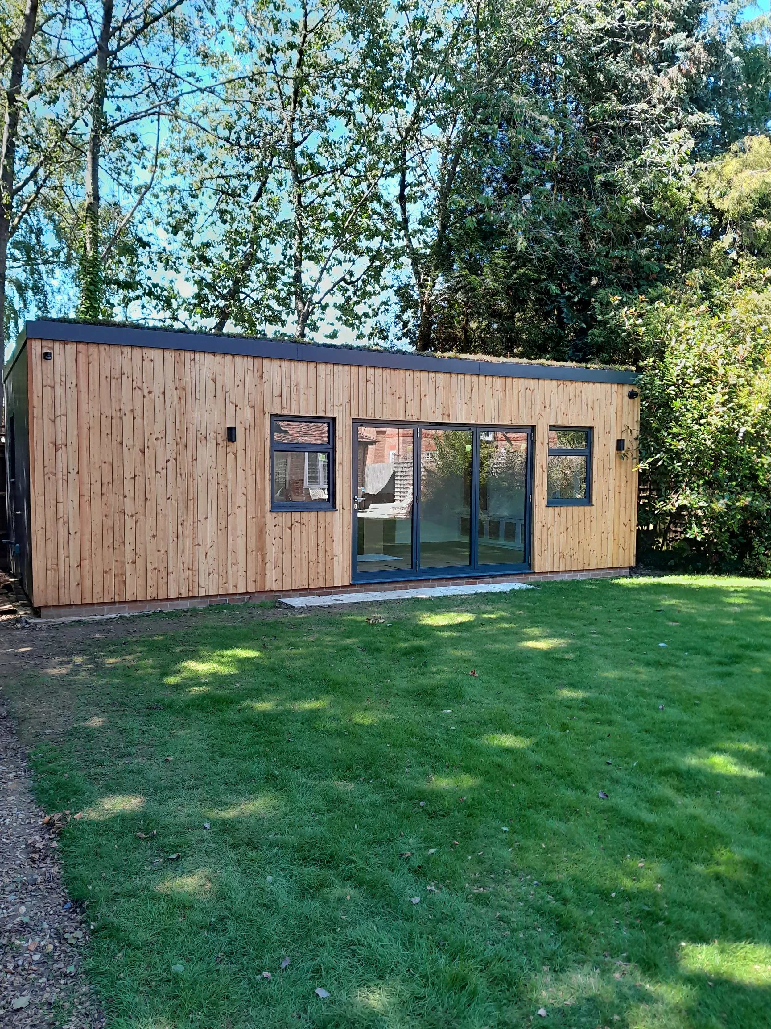 A small modern wooden building with black window and door frames, surrounded by a green lawn and tall trees.