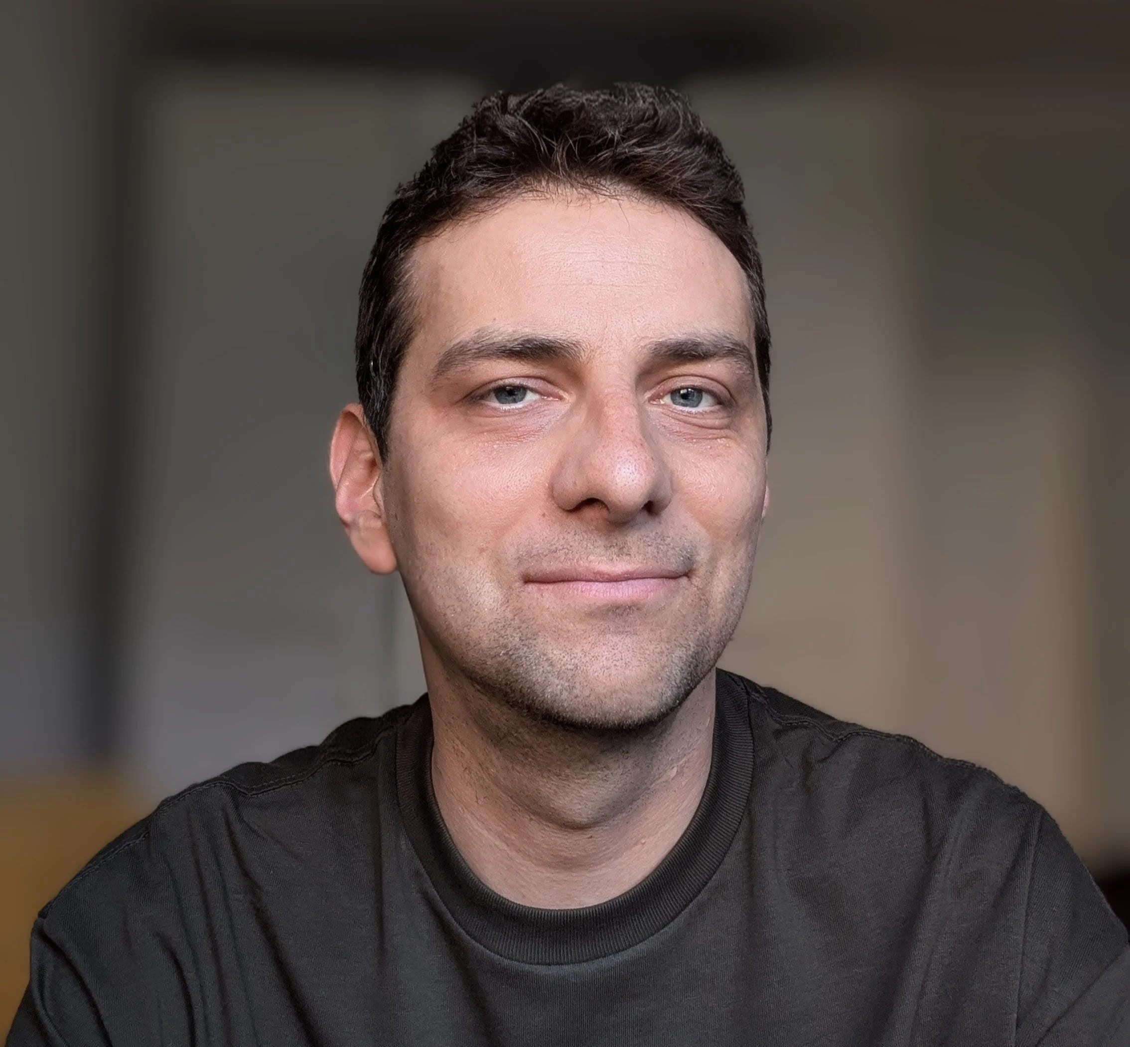 Close-up of a young man with dark hair, light blue eyes, wearing a black t-shirt.