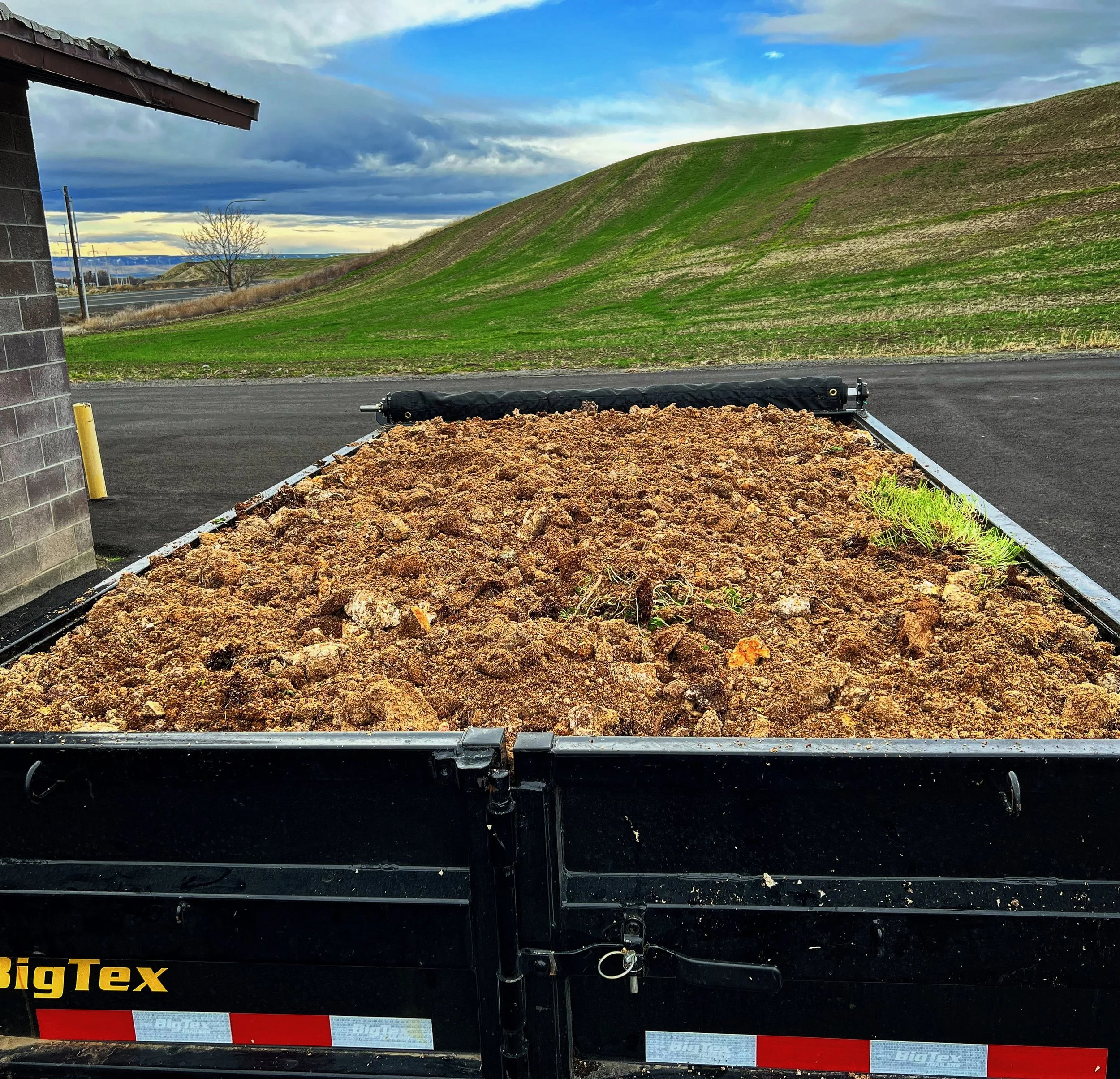 A trailer filled with dirt parked outside a building with a green hill and cloudy sky in the background.