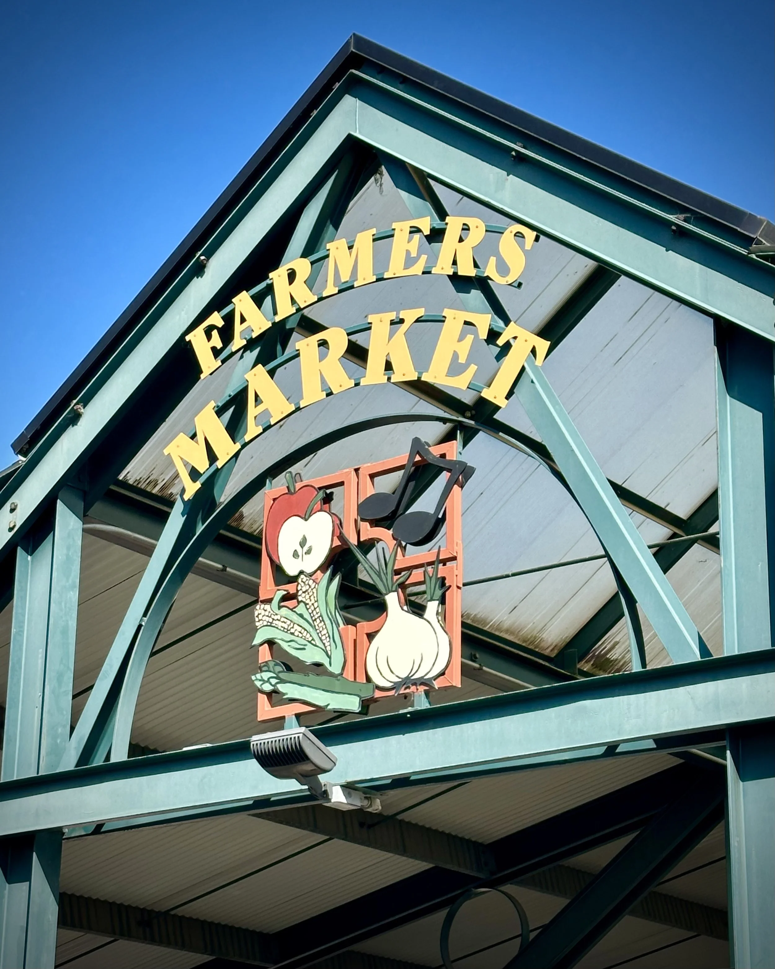 Sign for a farmers market with a decorative emblem featuring an apple, onion, and vegetable illustrations.