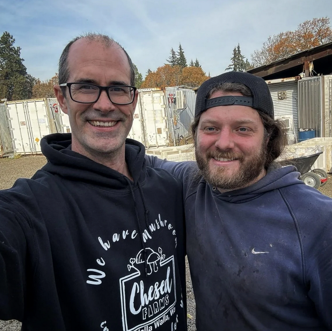 Two smiling men taking a selfie outdoors, with trailers and trees in the background, during what appears to be a cloudy day.