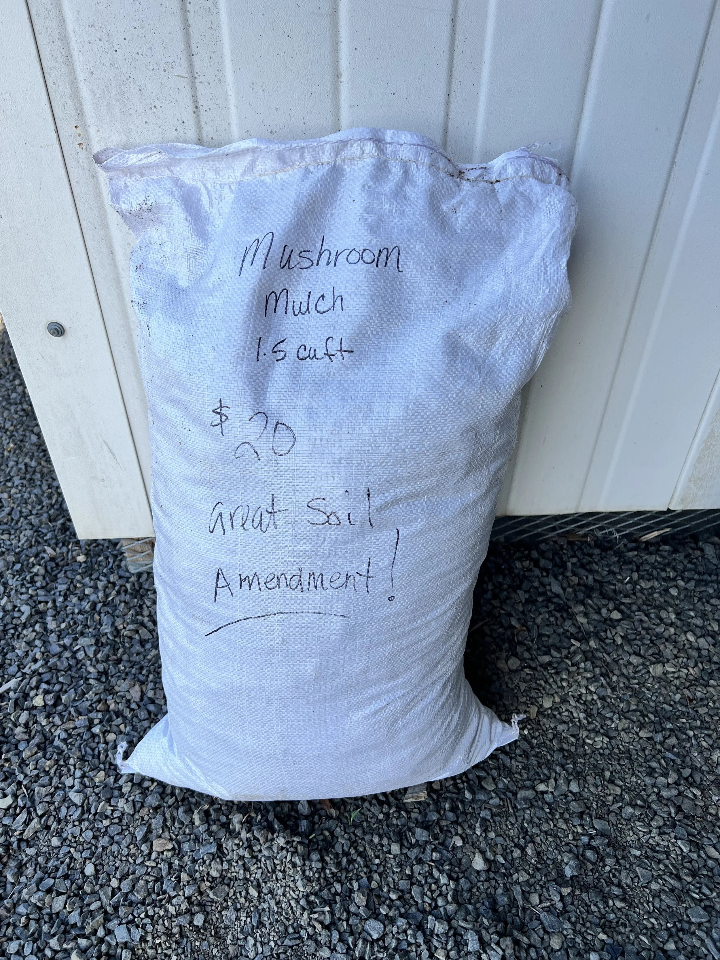 A white bag with handwritten notes about mushroom compost, priced at $20, lying on gravel outside next to a white wall.