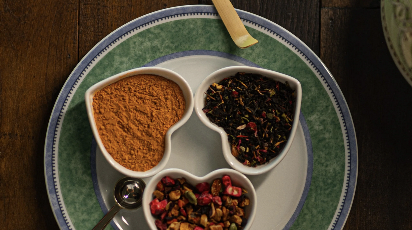 Three small bowls with different types of spices and dried herbs on a white plate, with a spoon and a green straw on a dark wooden table.