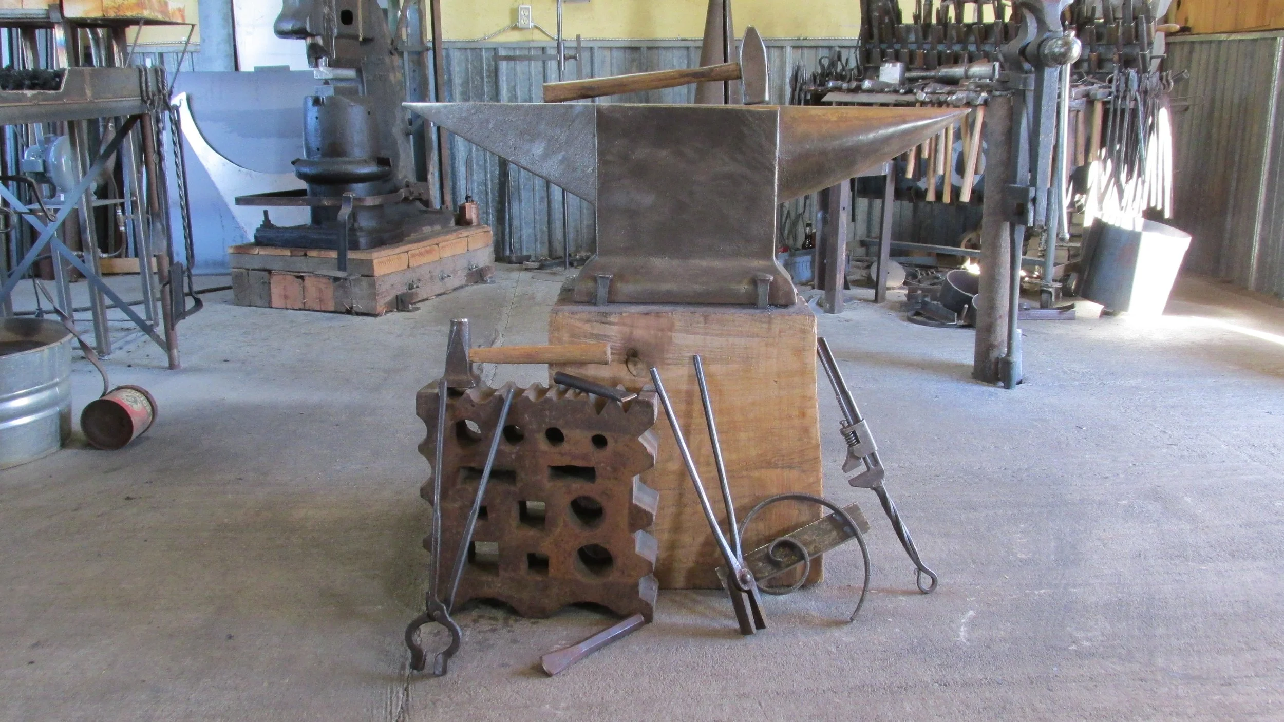 A blacksmith's forge with an anvil on a wooden block, surrounded by blacksmithing tools such as tongs and hammers, inside a workshop with metalwork equipment and tools hanging on the walls.