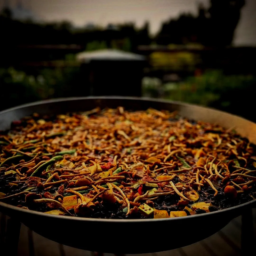 A large pan of stir-fried noodles with vegetables, set outdoors with a blurred background of a rooftop garden or balcony.