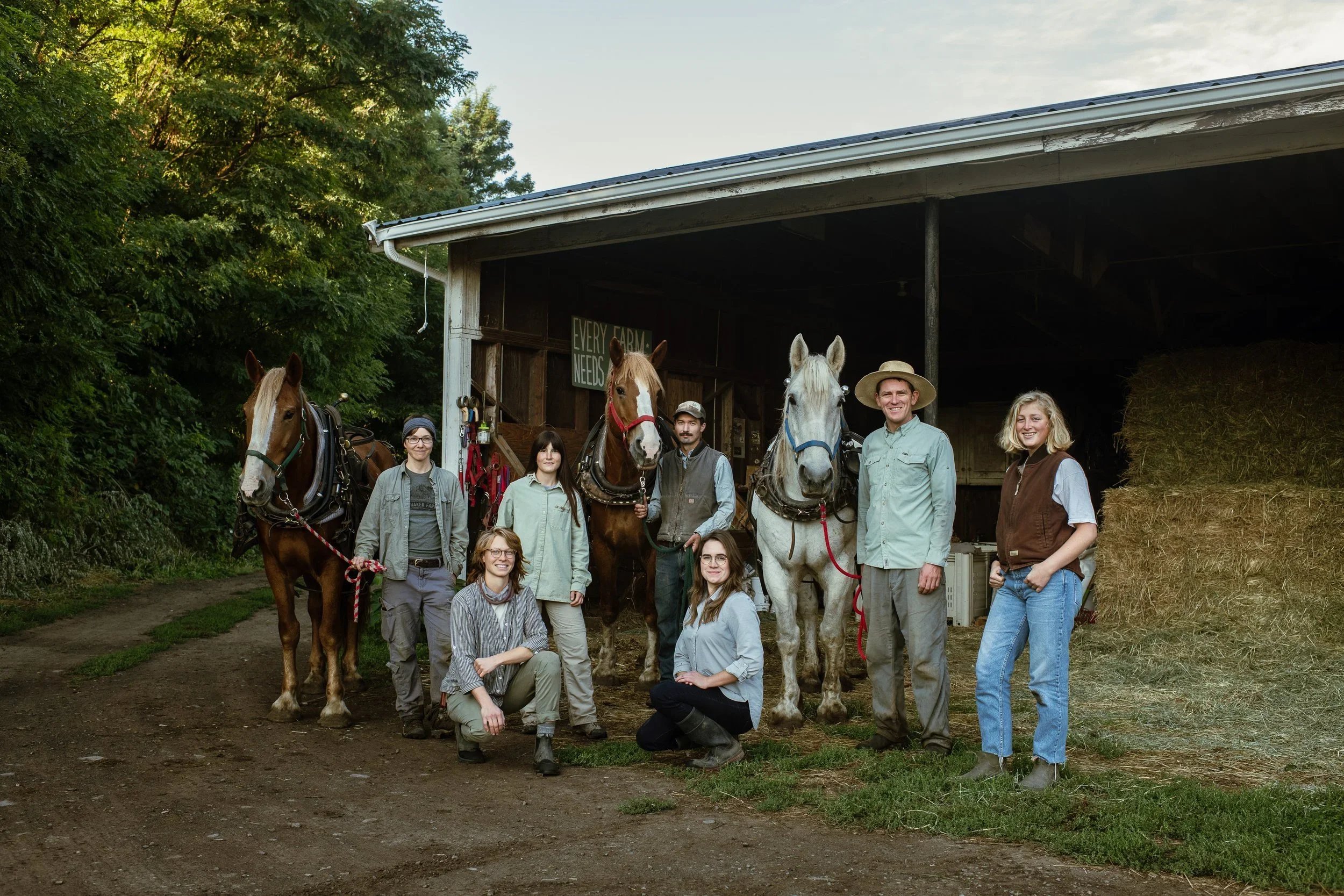 Group of nine people standing and kneeling in front of a barn with four horses. Some people are smiling, and there is hay stacked on the right side of the barn. The scene appears to be at a farm or ranch.