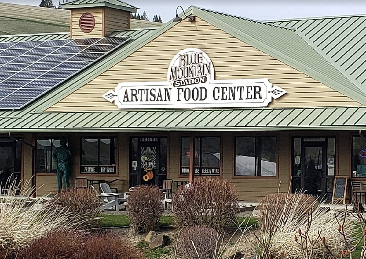 Front view of a building with a sign that reads 'Blue Mountain Station Artisan Food Center'. The building has a green metal roof with solar panels, tan siding, and several windows. There are outdoor chairs and bushes in the foreground.