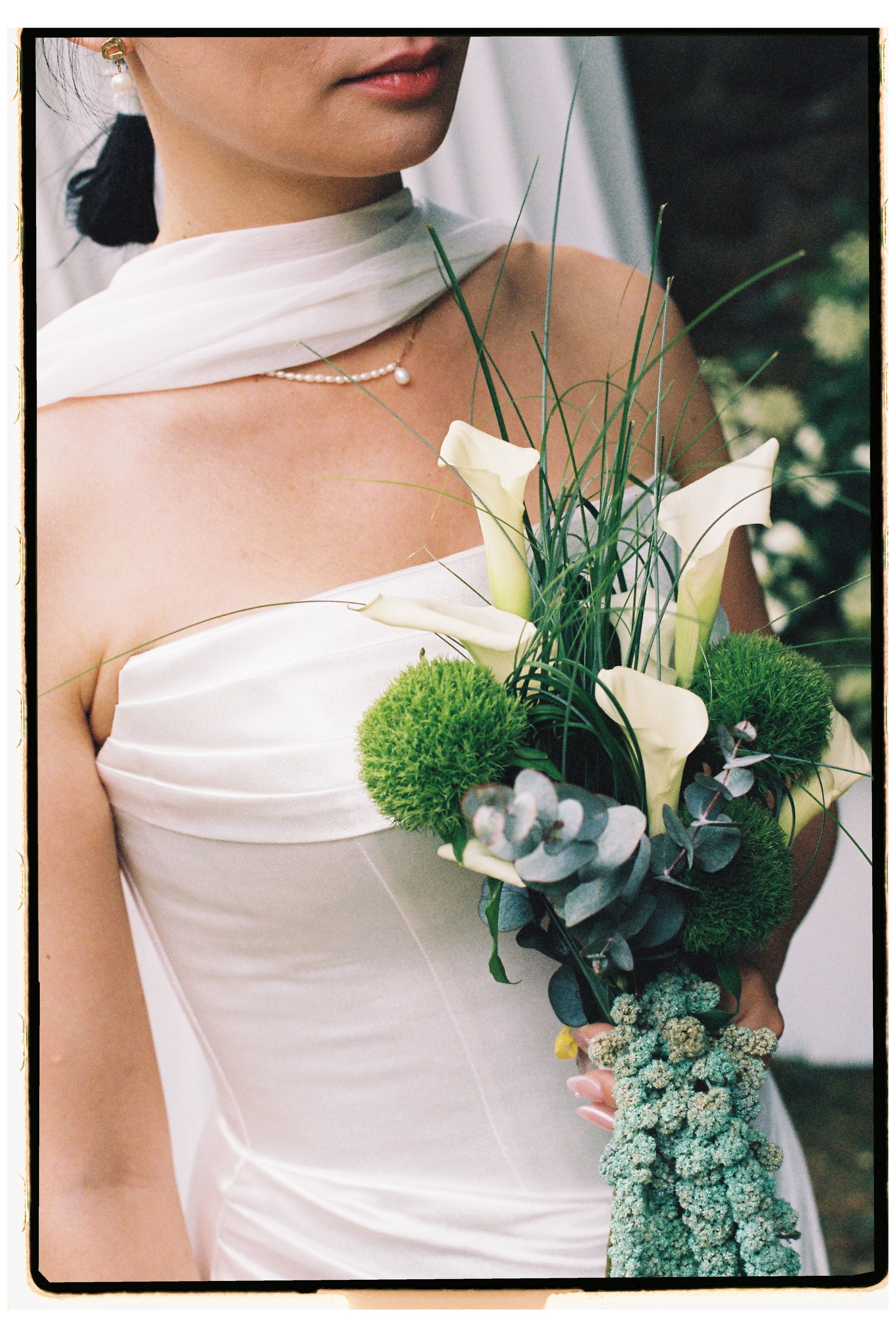 A woman in a white dress holding a bouquet of white and green flowers, including calla lilies, green moss, eucalyptus, and other greenery, with a pearl necklace and a pearl earring. The photo is a close-up of her upper body and face.