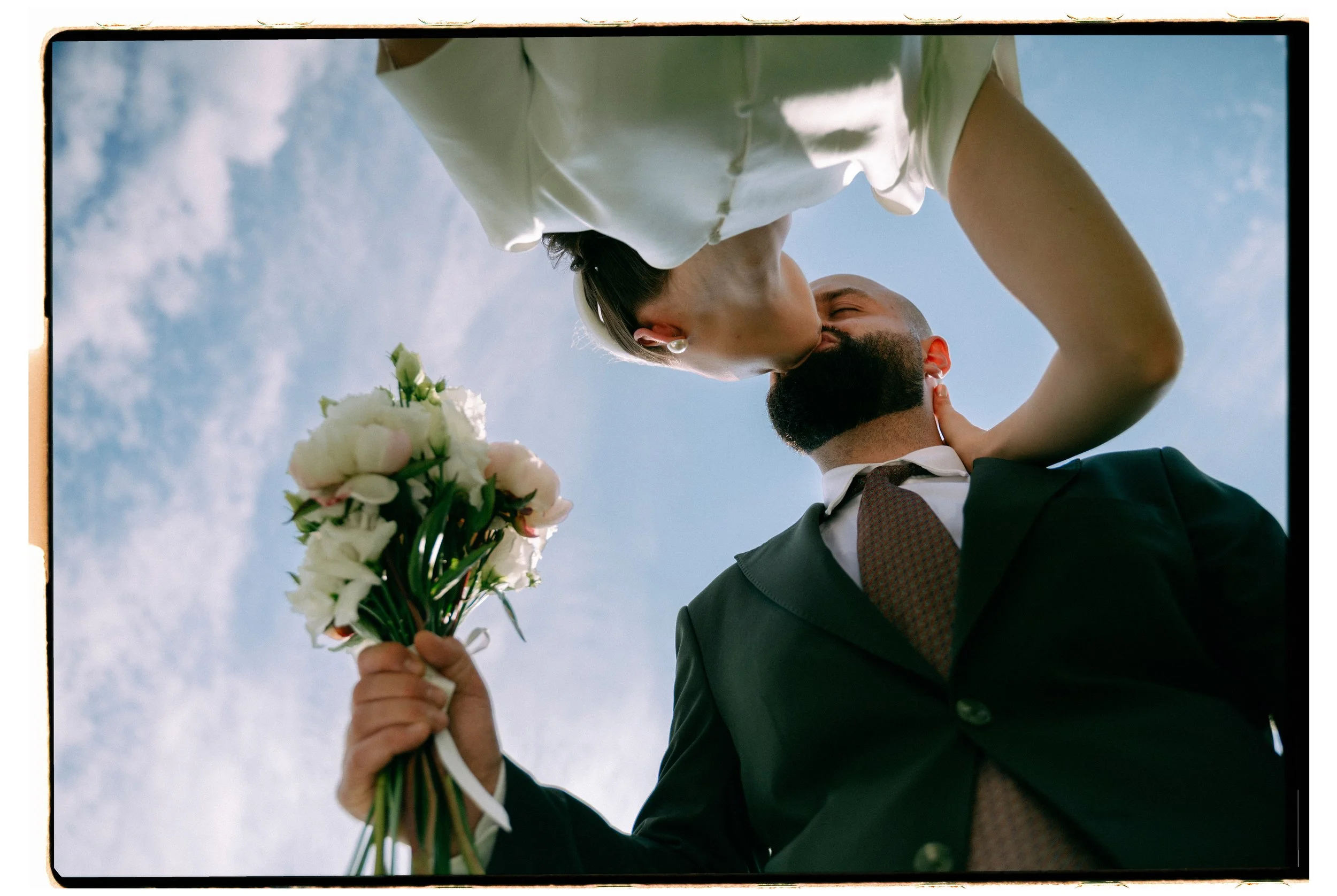A couple kissing outdoors, with one holding a bouquet of white flowers against a blue sky with light clouds.