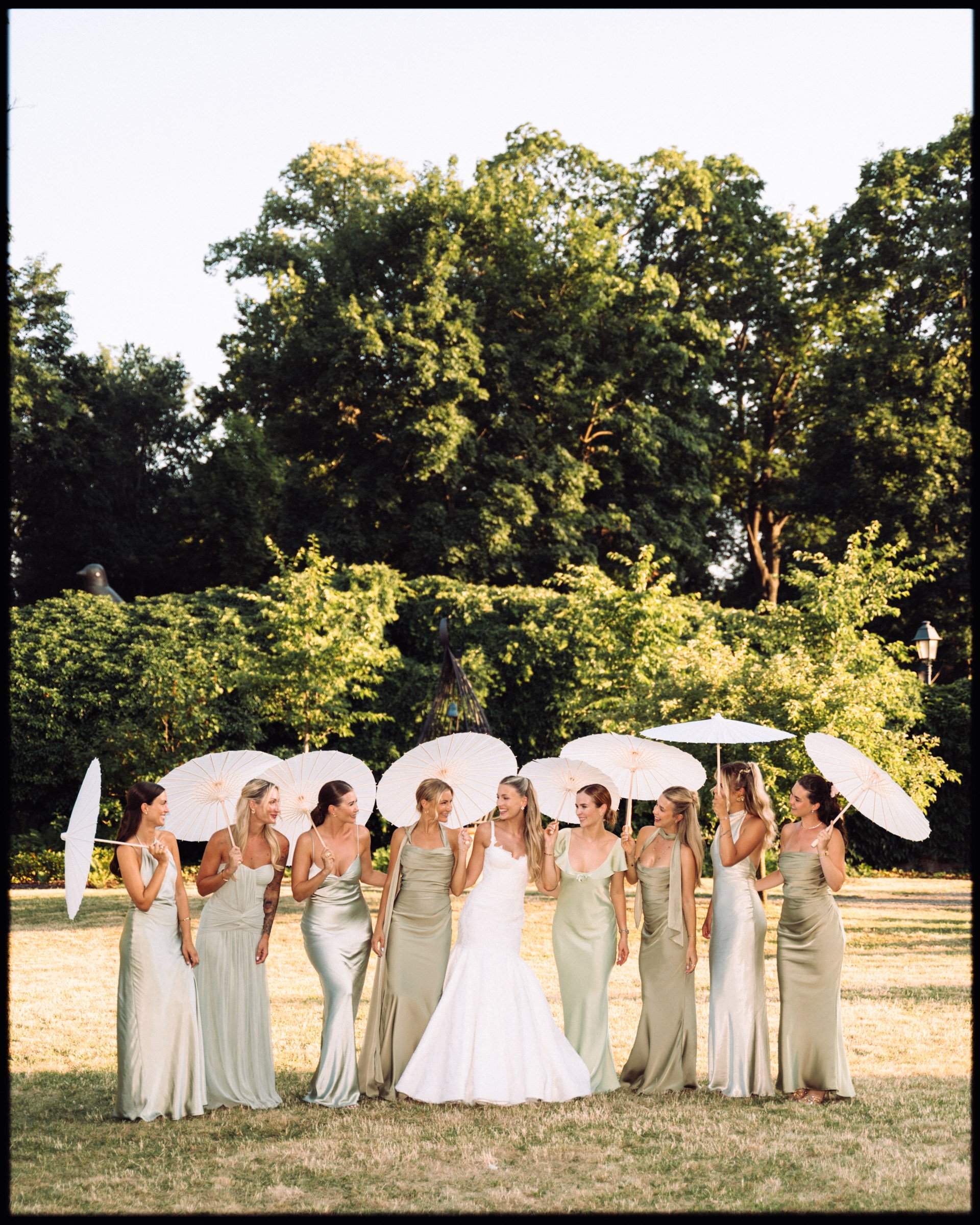A bride and eight bridesmaids standing outdoors on a grassy area with trees in the background, holding white parasols. The bridesmaids are dressed in matching light-colored dresses, and the bride is wearing a white wedding gown.