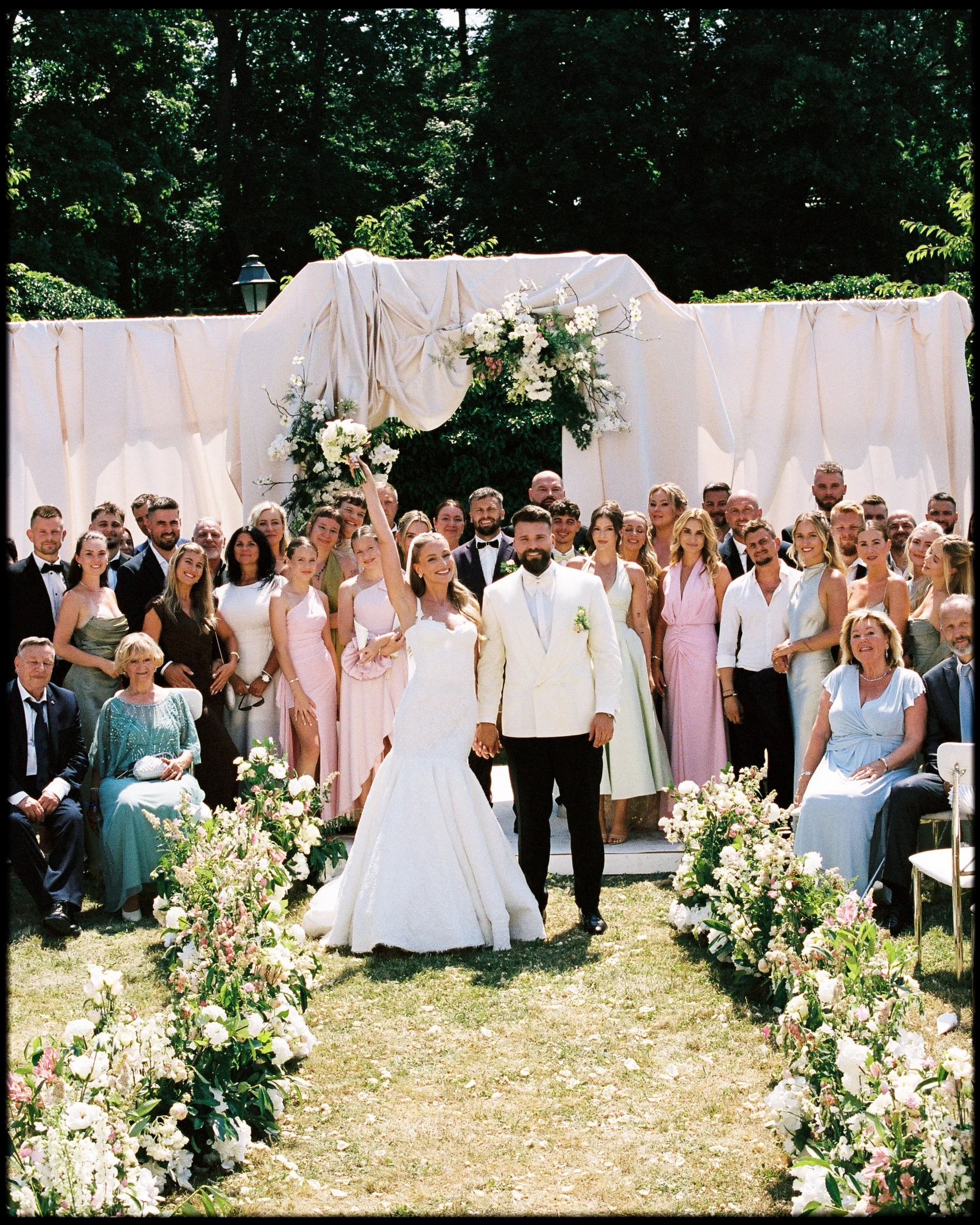 A large group of people dressed in formal attire at an outdoor wedding ceremony, with a bride and groom in the center holding hands, surrounded by floral arrangements and greenery.