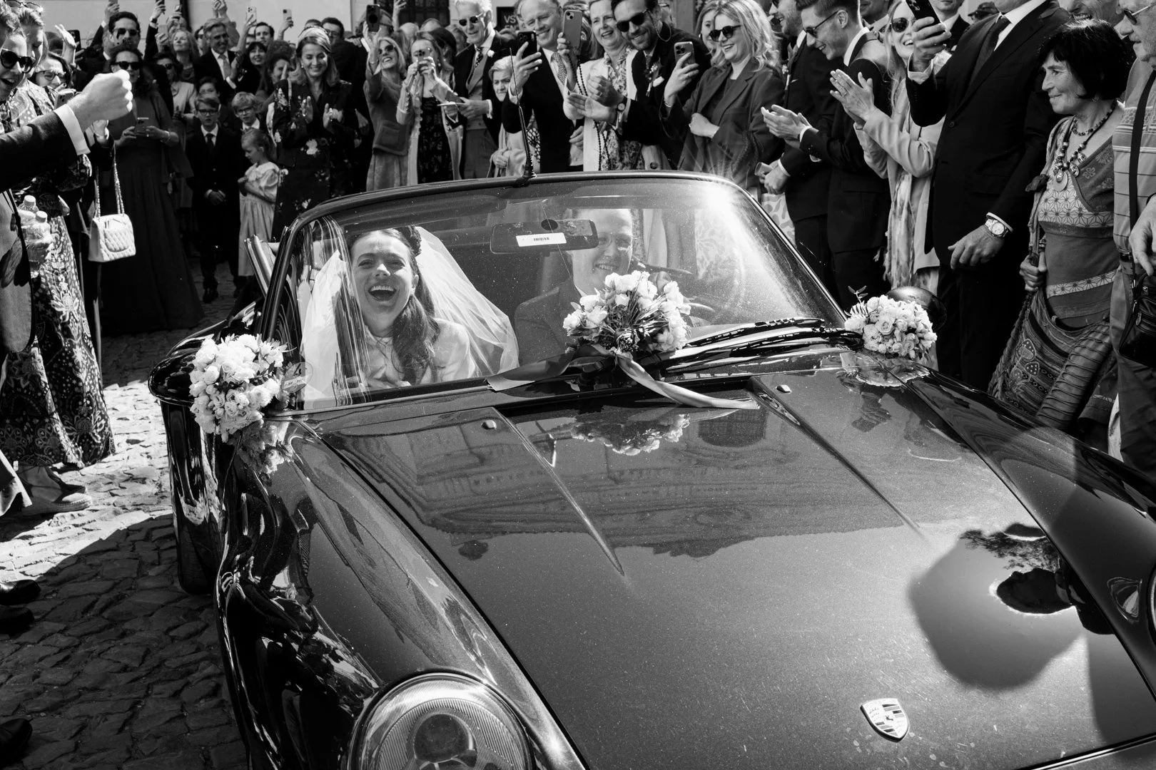 A couple dressed as newlyweds in a vintage convertible car decorated with flowers, surrounded by a crowd of happy, taking photos and cheering, during a wedding celebration.