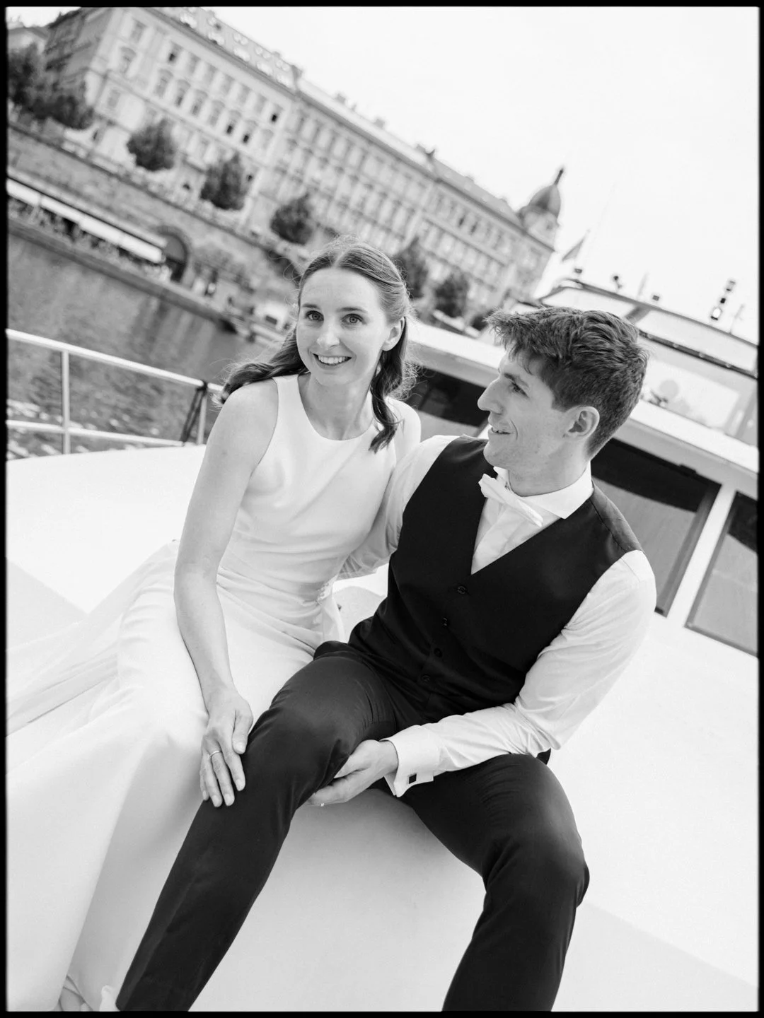 A black and white photo of a smiling woman in a white dress and a man in a vest and white shirt sitting on a boat with a cityscape and water in the background.