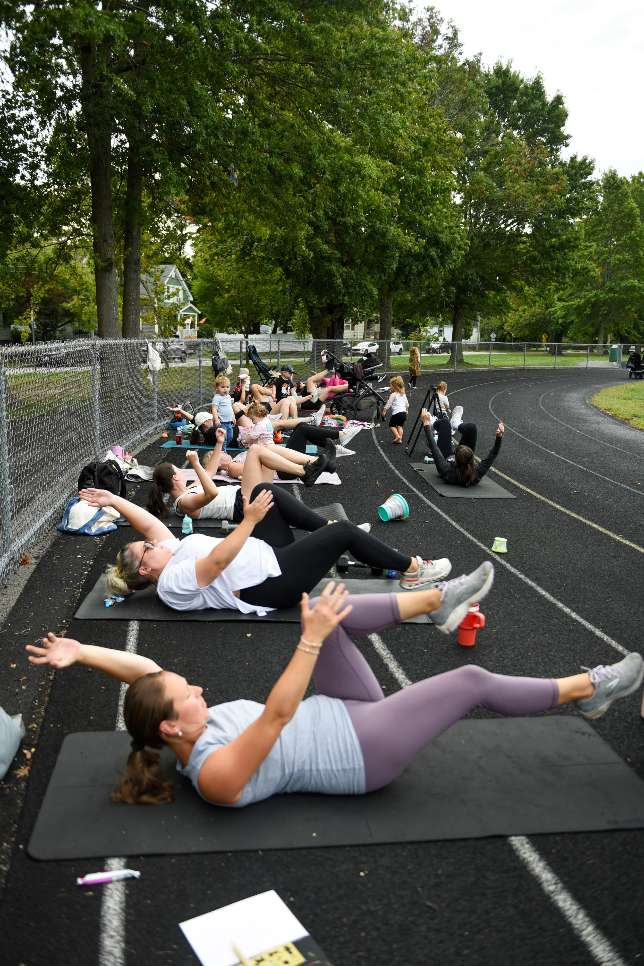 Group of SouthCoast moms and children participating in an iStroll SouthCoast fitness class at Cushman Park in Fairhaven, lying on exercise mats and doing stretching exercises, with some kids playing nearby.
