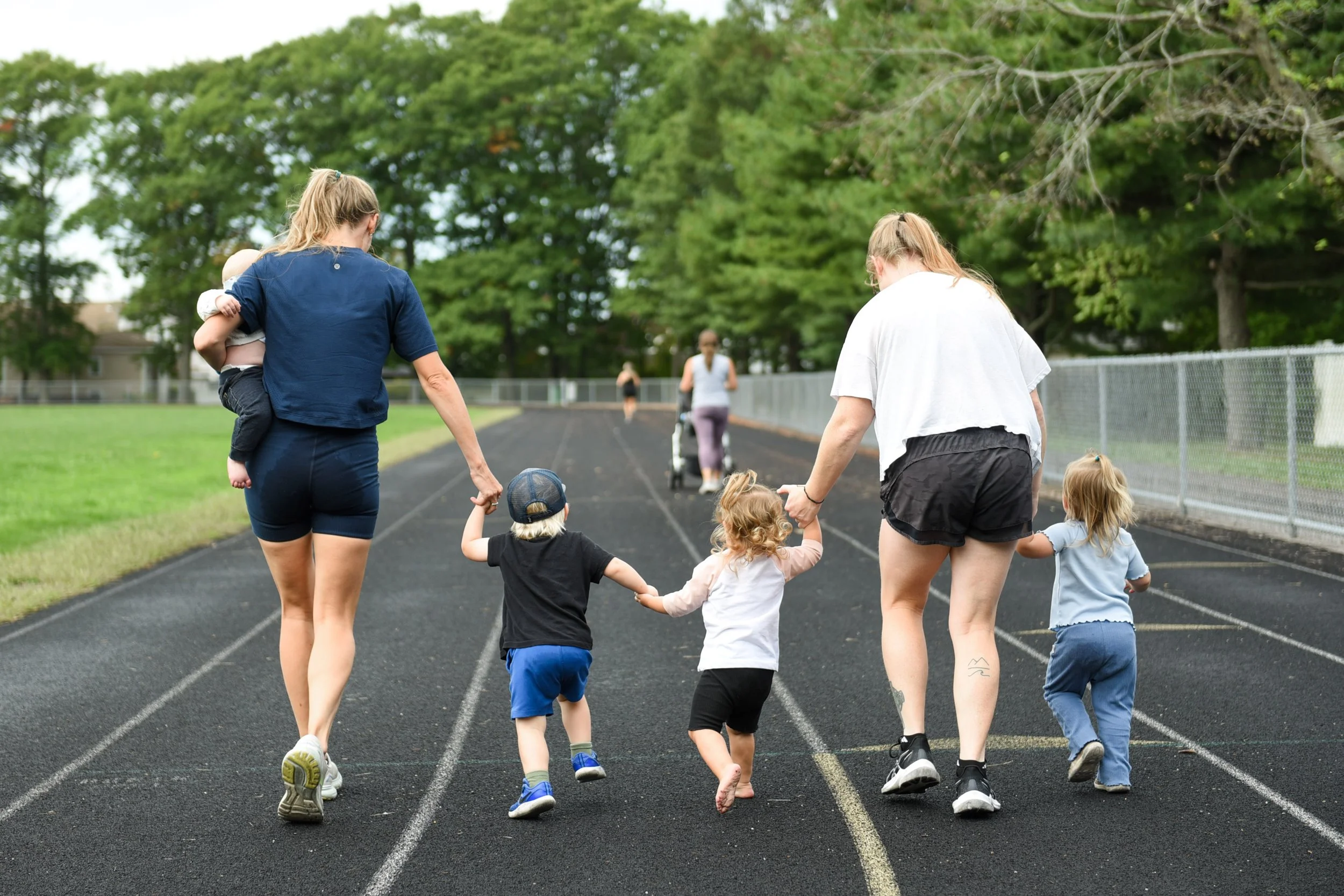 Two SouthCoast moms, each holding the hand of a young child, walking on the track at Cushman park in Fairhaven at iStroll Southcoast with lush green trees in the background.