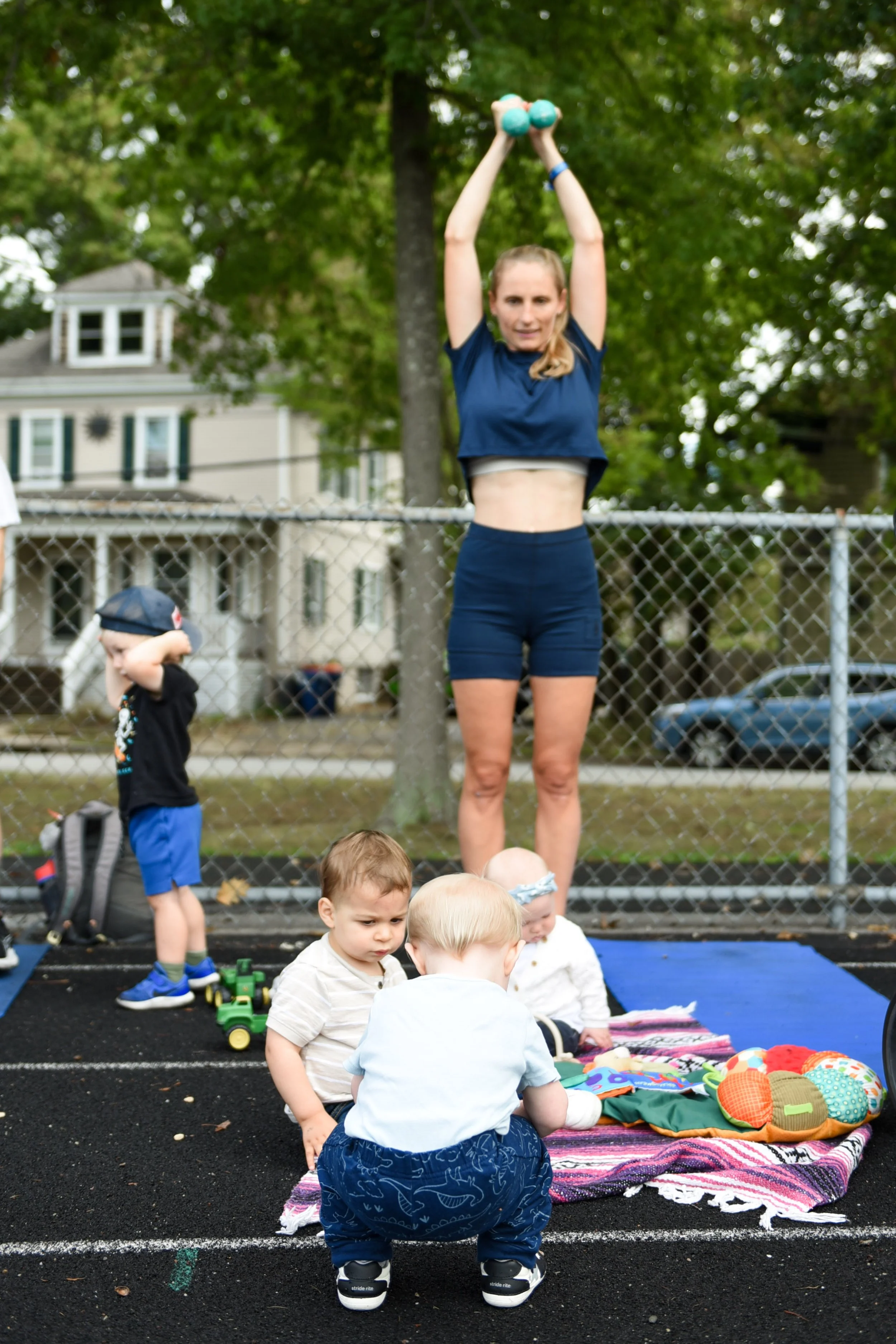 A woman exercising outdoors with a light blue dumbbell overhead, children playing nearby on a playground surface with colorful toys, and a house and trees in the background at iStroll SouthCoast