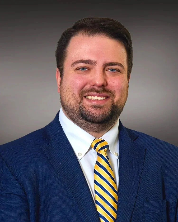 Professional headshot of a man in a navy blue suit and yellow striped tie, smiling against a gray blurred background.