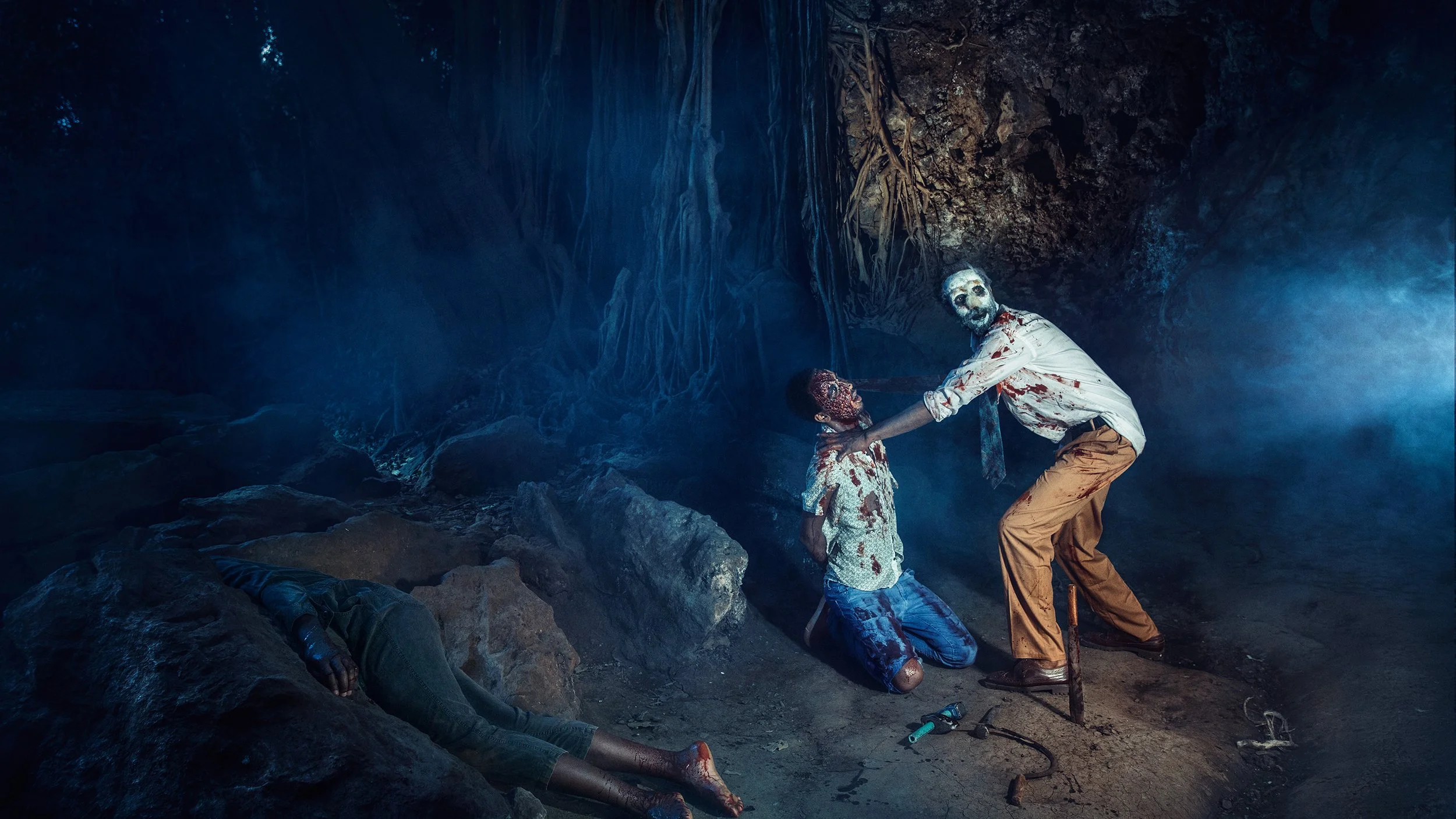 A person with a creepy, zombie-like mask and bloodstained clothing holds the arm of a kneeling young boy with a mask and bloodstained clothes inside a dark, cave-like setting. Another person is lying on the ground in the foreground.