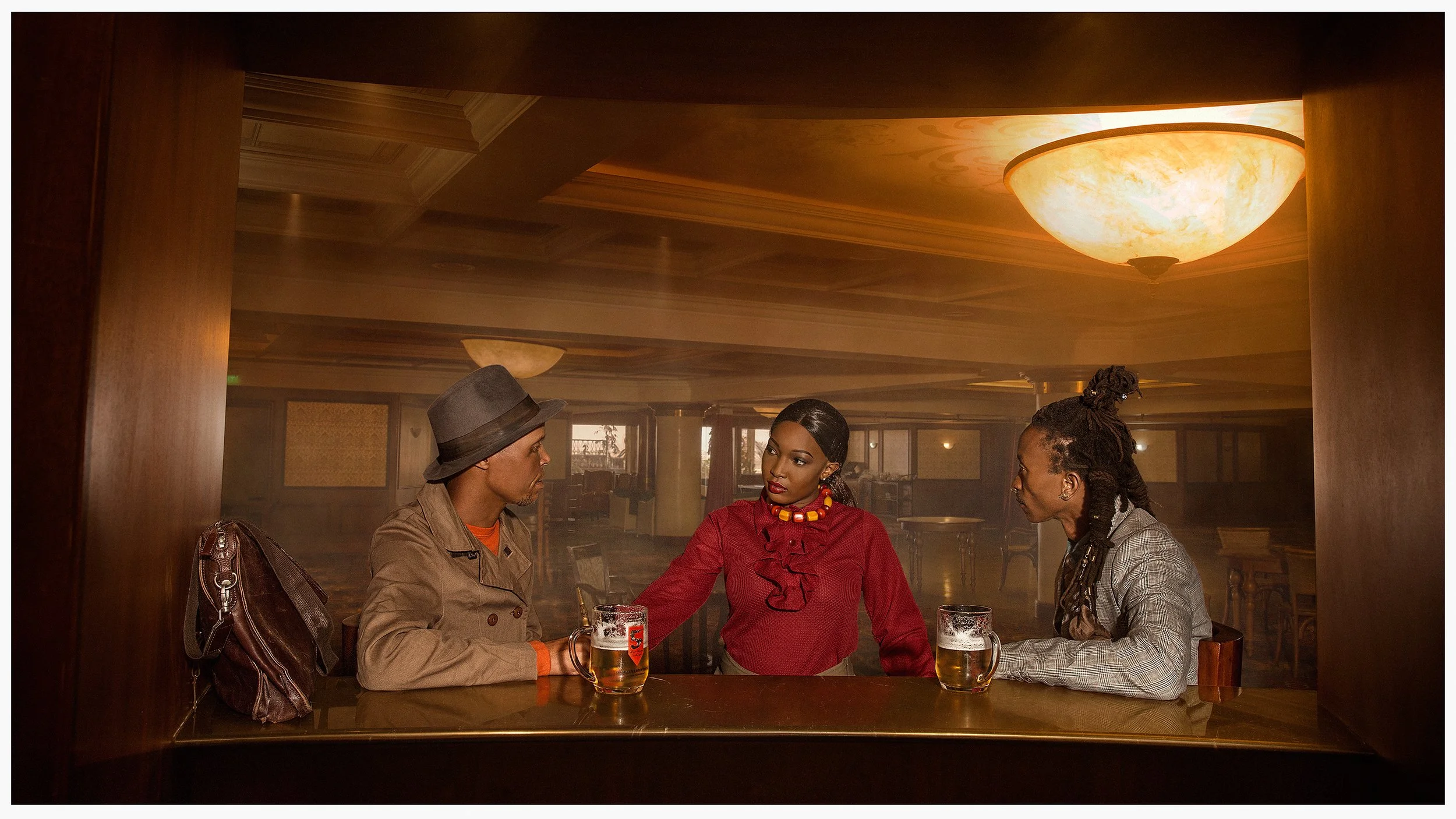 Three people sitting at a bar or restaurant table, engaged in conversation. A man with light skin, wearing a hat and tan coat, is on the left. A woman with dark skin, wearing a red blouse and colorful necklace, is in the middle. A man with dark skin, wearing a gray checked blazer, is on the right. Two glasses of beer are on the table. The background shows a warmly lit, upscale interior with ceiling lights and wood paneling.