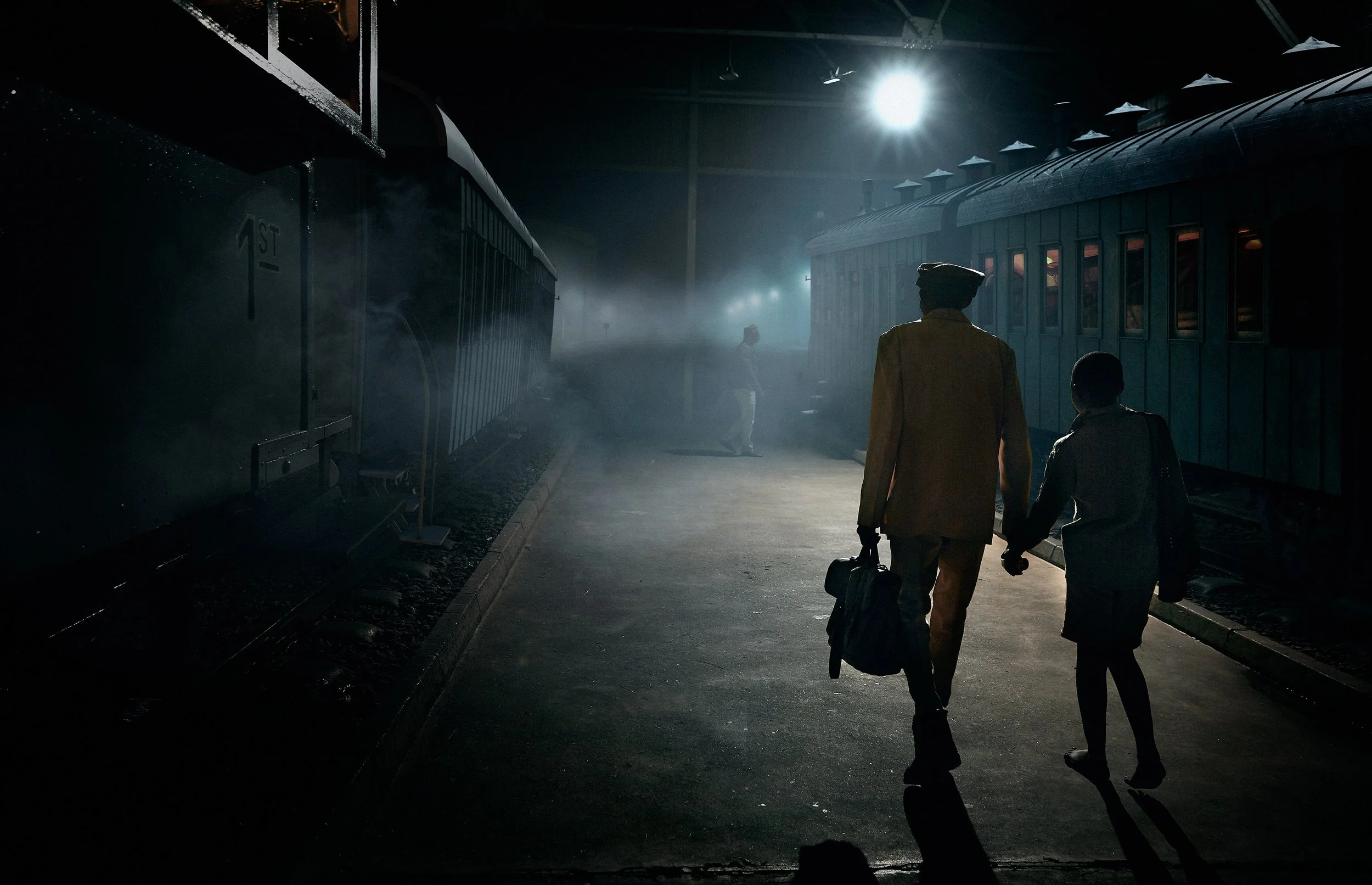 A train station at night with two men walking away from the camera, carrying luggage, and a third person standing near the train, illuminated by a bright overhead light.