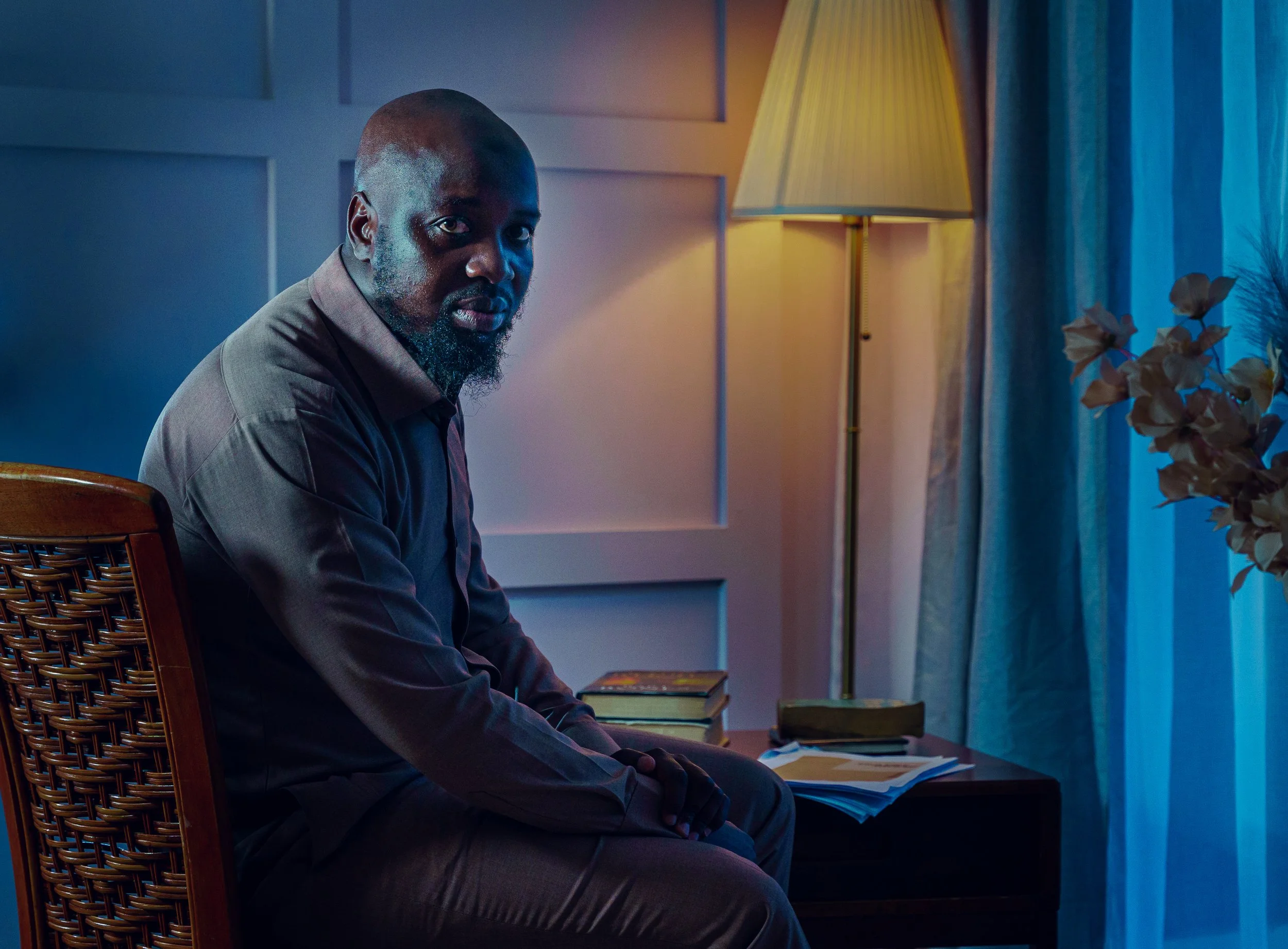 A man with a beard and shaved head sitting in a dimly lit room, illuminated by warm and cool light, with a lamp, flowers, and books visible on the side.