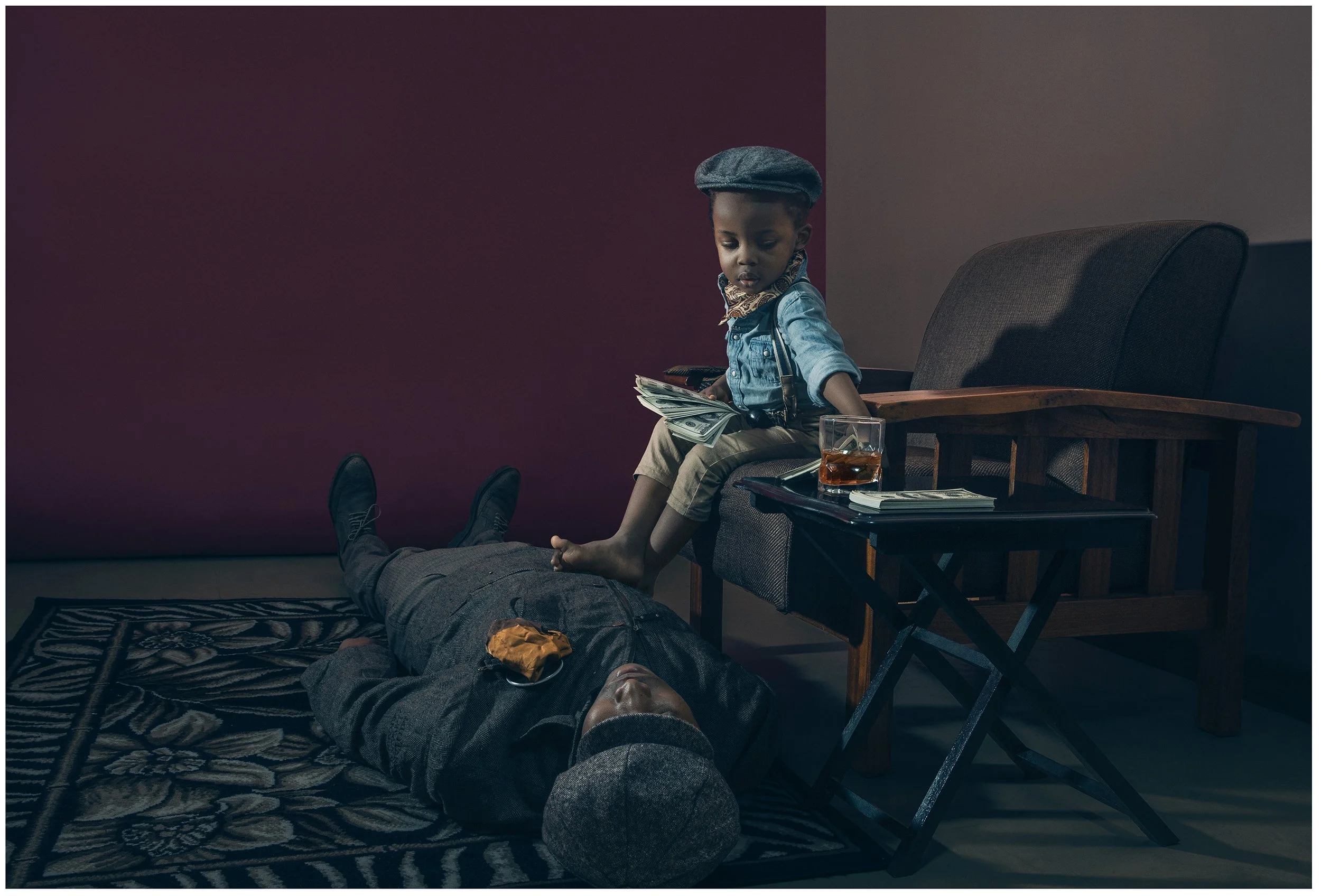 A young boy with a dark hairstyle, wearing a denim shirt, tan pants, a gray beret, and a patterned scarf, sitting on a vintage armchair holding a stack of cash. A glass of whiskey, a remote, a notebook, and a smartphone are on a small table nearby. A person with dark pants and shoes is lying on a patterned rug on the floor, with their head and feet visible. The background features a two-tone wall with maroon and beige colors.