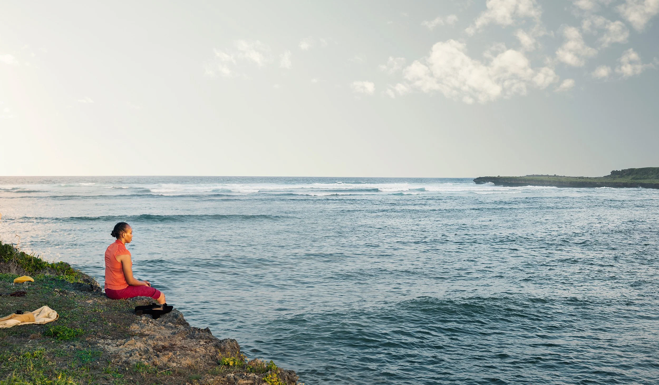 A woman sitting cross-legged and meditating on a rocky shoreline by the ocean, with waves in the water and a cloudy sky above.