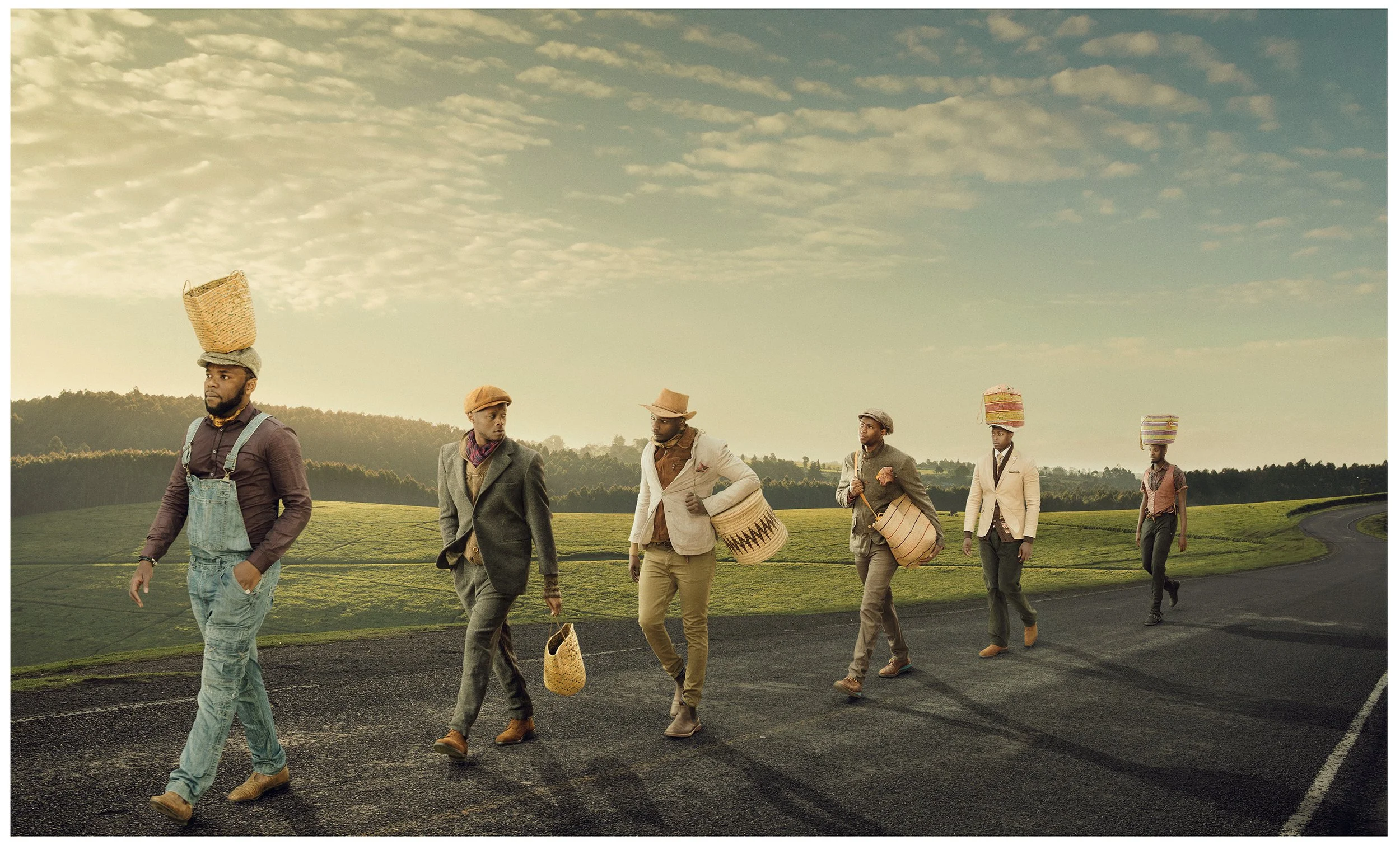 A group of six men dressed in vintage attire walking across a countryside landscape with rolling green hills under a partly cloudy sky, carrying woven baskets and wearing hats.