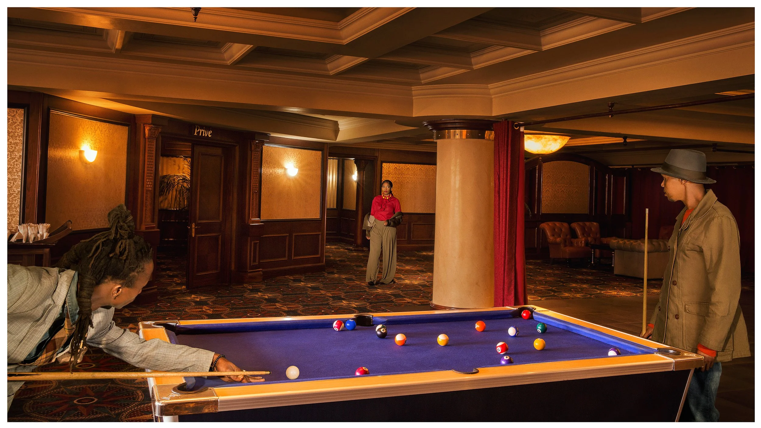 People playing pool in a dimly lit room with wood-paneled walls and a patterned carpet, with a woman standing in the background near a doorway labeled 'Prive'.