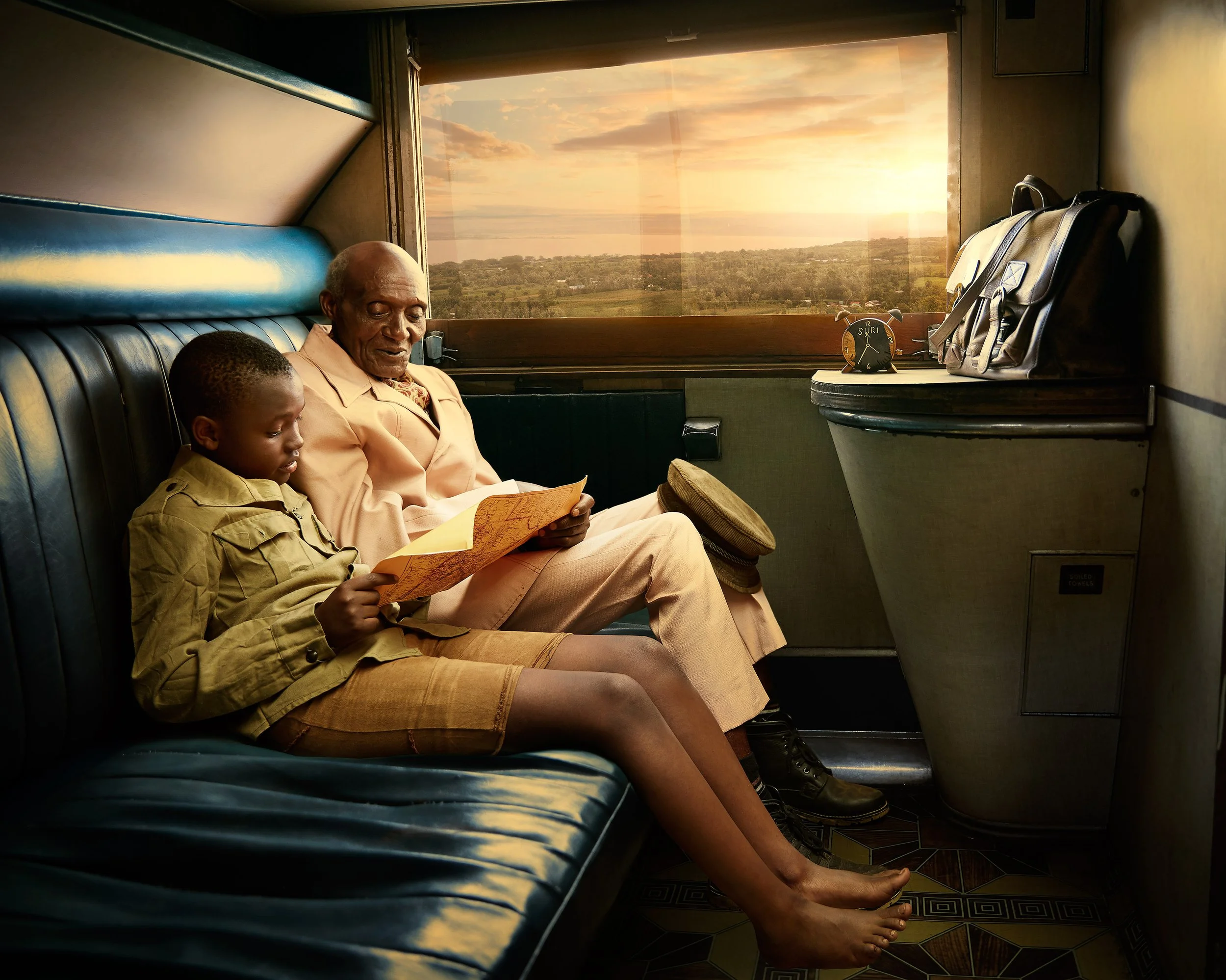 A man and a young boy sitting on a dark blue train seat, reading a book together. The window behind them shows a scenic view of a landscape with trees and a sunset sky.