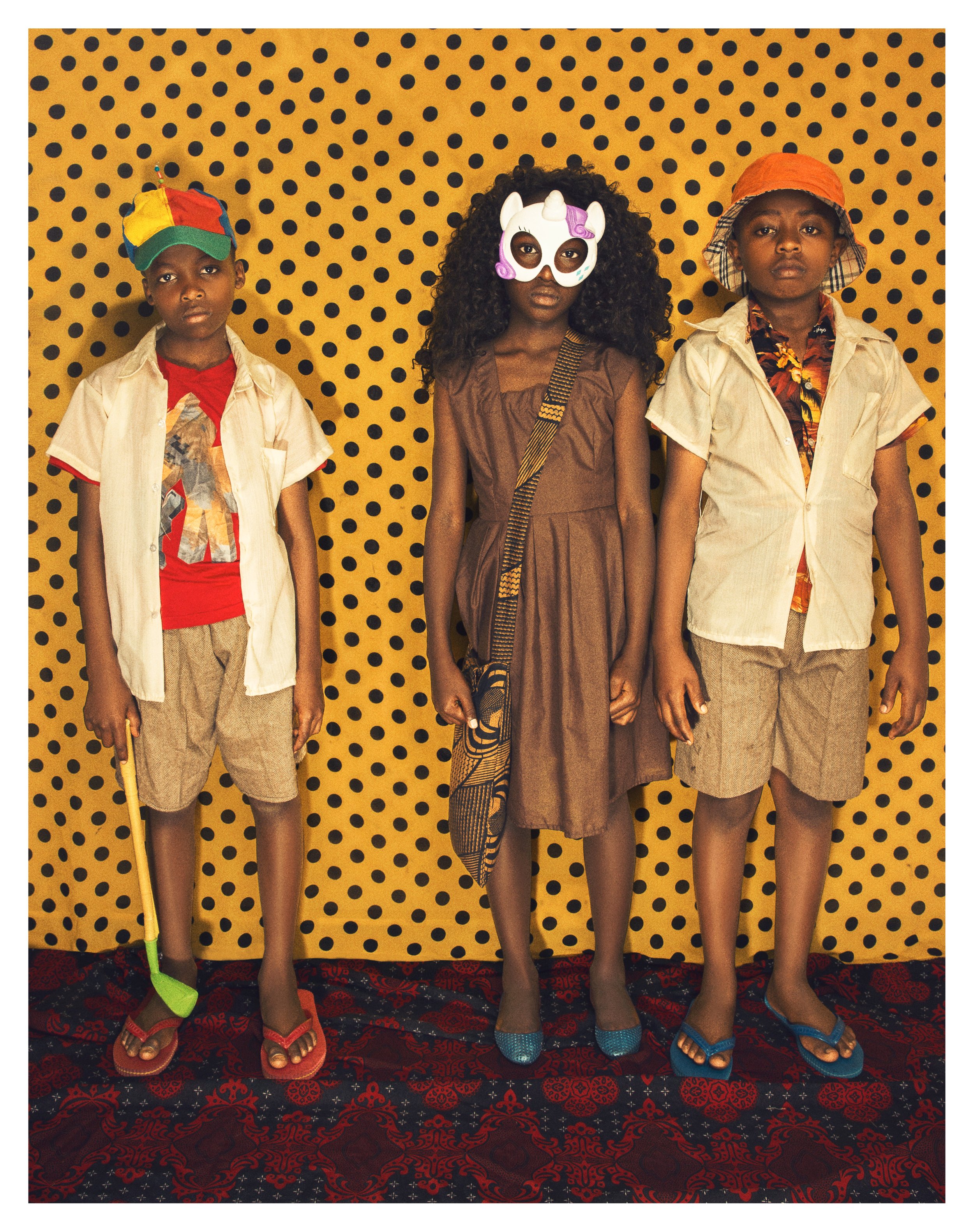 Three children stand in front of a yellow background with black polka dots, dressed in casual summer clothes and wearing colorful hats and masks, with a red and black patterned carpet beneath them.