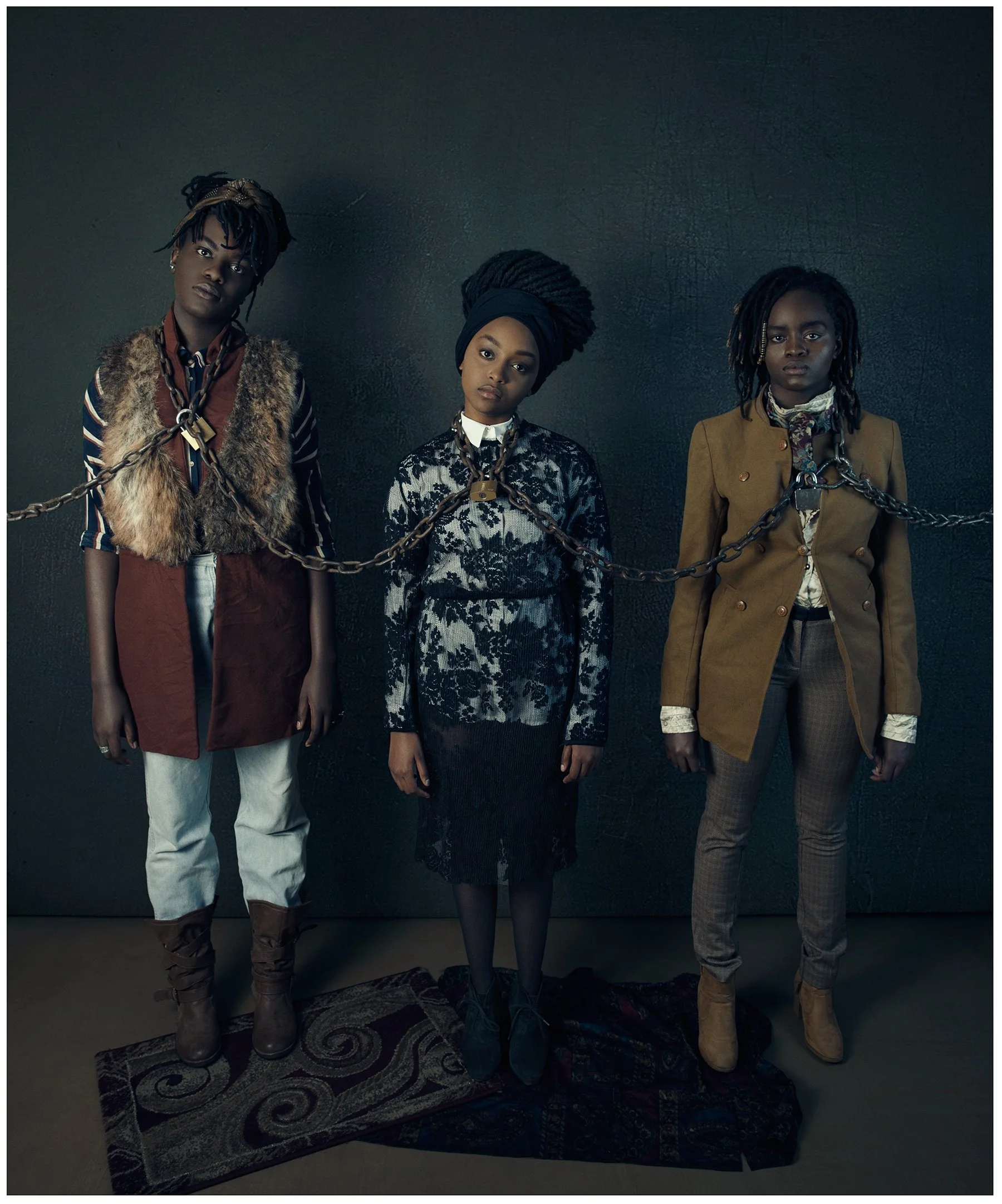 Three women standing against a dark wall, each wearing brown chains around their necks and shoulders, with serious expressions. The woman on the left has dreadlocks and wears a fur vest over a striped shirt, white pants, and brown boots. The woman in the middle has her hair in an updo with a headband, and wears a black and white lace dress. The woman on the right has dreadlocks and wears a tan blazer, patterned shirt, and tan boots.