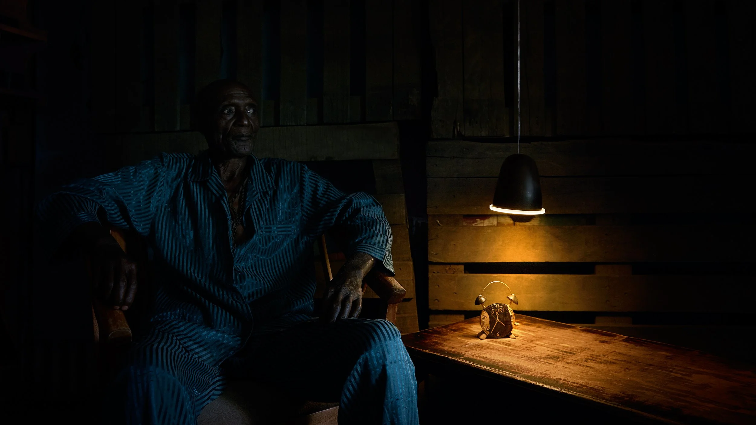 An elderly man sitting in a dimly lit room, illuminated by a single hanging lamp and a small clock on a wooden table nearby.