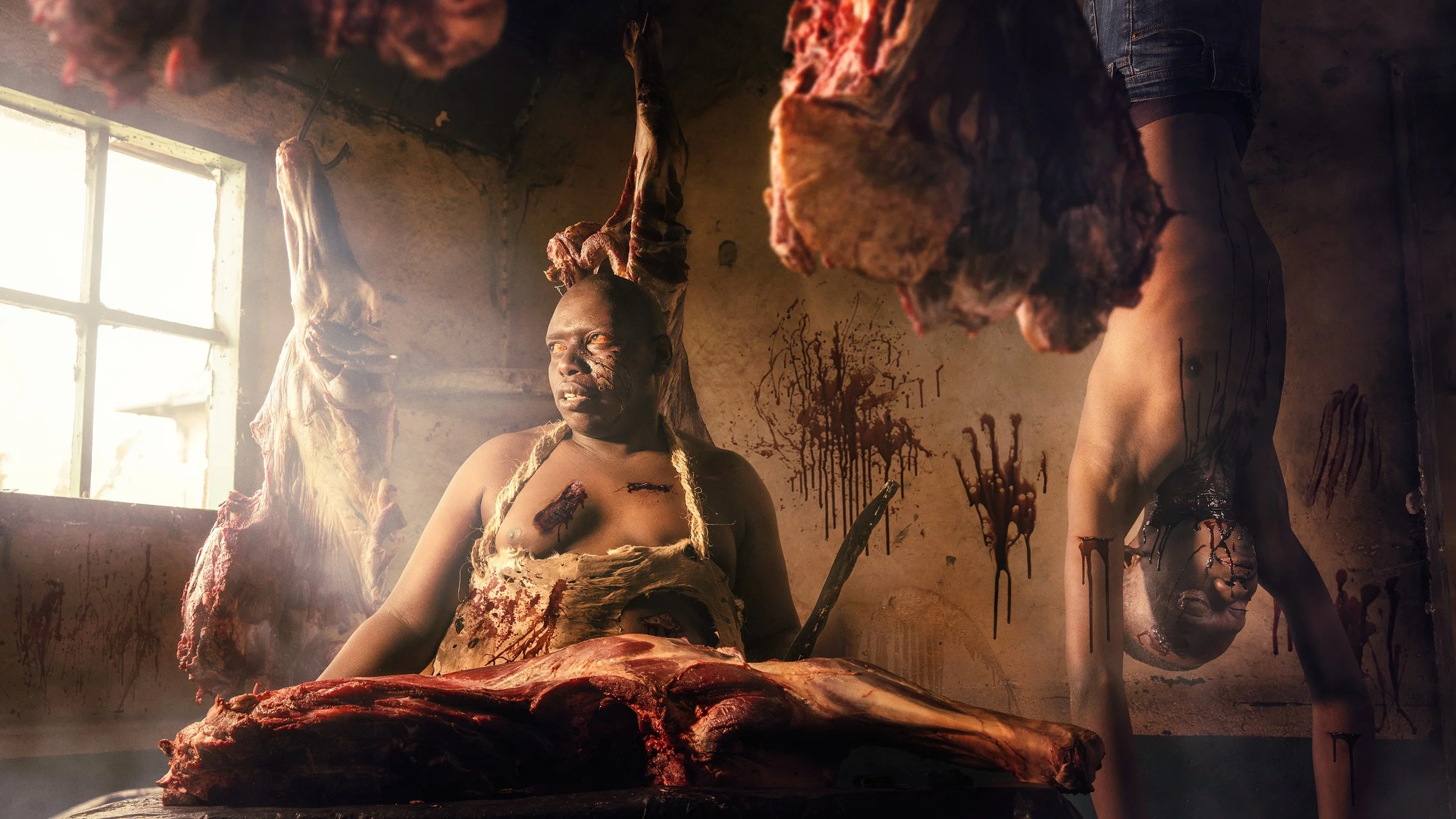 Two people with disheveled hair and bodies covered in blood and wounds, standing in a dimly lit room with blood splatters on the wall, surrounded by hanging carcasses of animals.