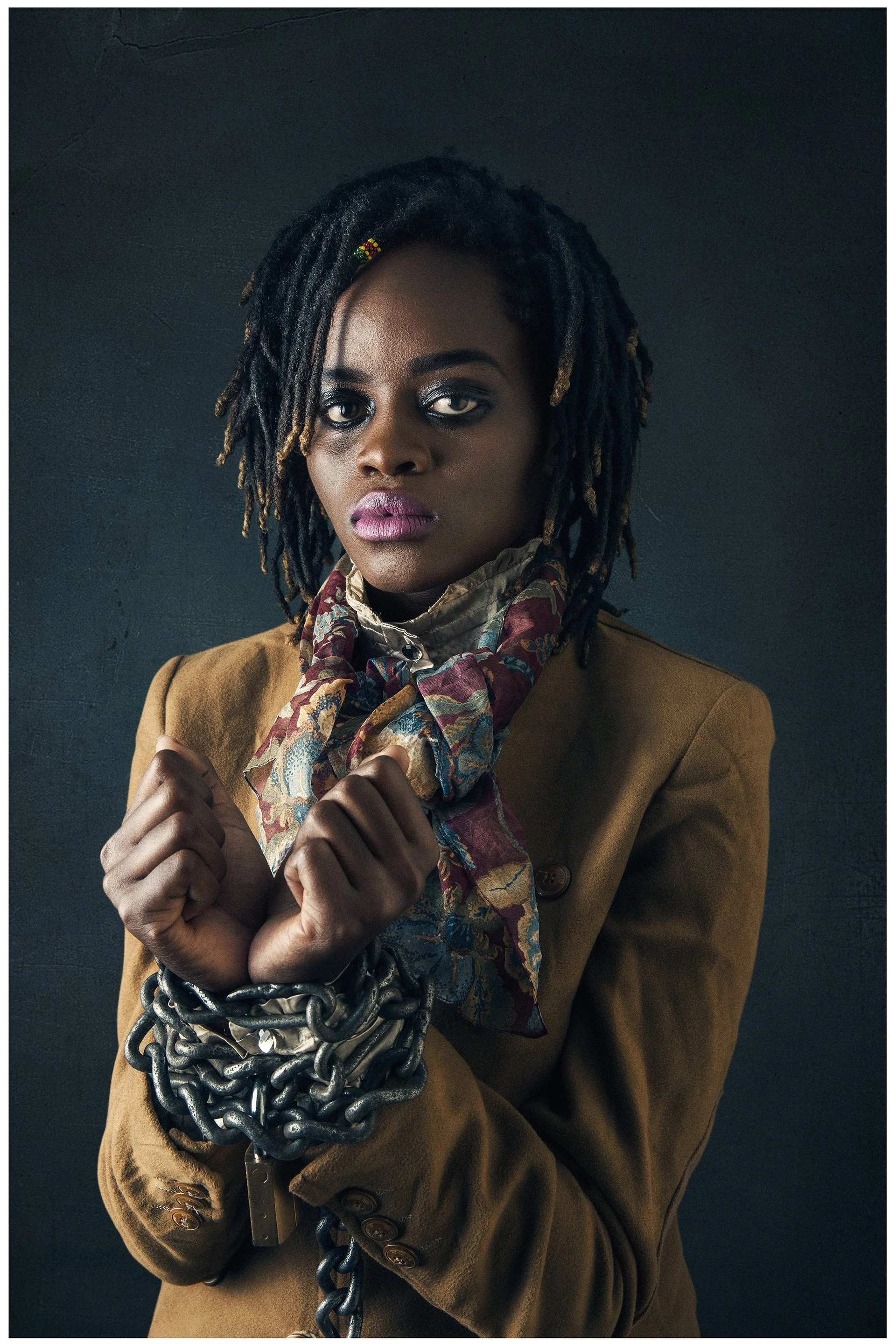 A woman with dark skin and dreadlocks wearing a brown blazer and a colorful scarf, holding chains and metal cuffs, with a serious expression against a dark background.