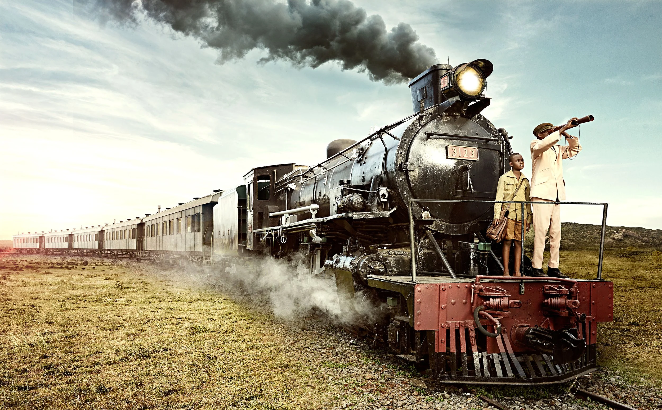 A vintage steam train with black smoke emitting from its chimney, moving across a grassy landscape under a cloudy sky. Two people stand on the front platform: a man using a telescope and a young boy beside him, both dressed in vintage clothing.