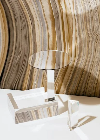 Decorative art piece consisting of a large wall with marbled brown and beige patterns and three clear geometric glass sculptures on a white surface.
