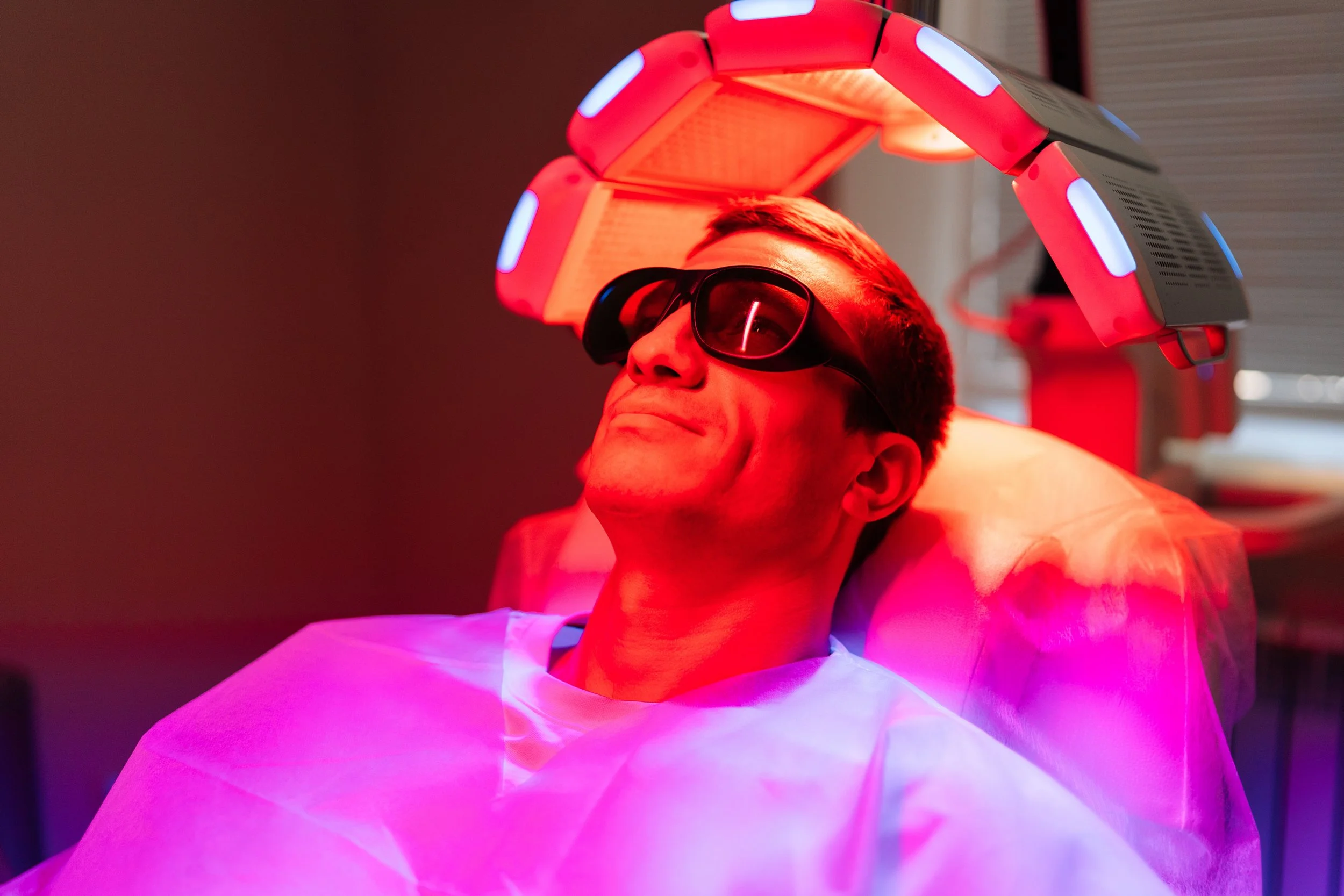 A man lying in a medical or spa setting, wearing glasses, with a reddening ultraviolet light therapy lamp over his head. The lighting is red and pink, creating a calming atmosphere.