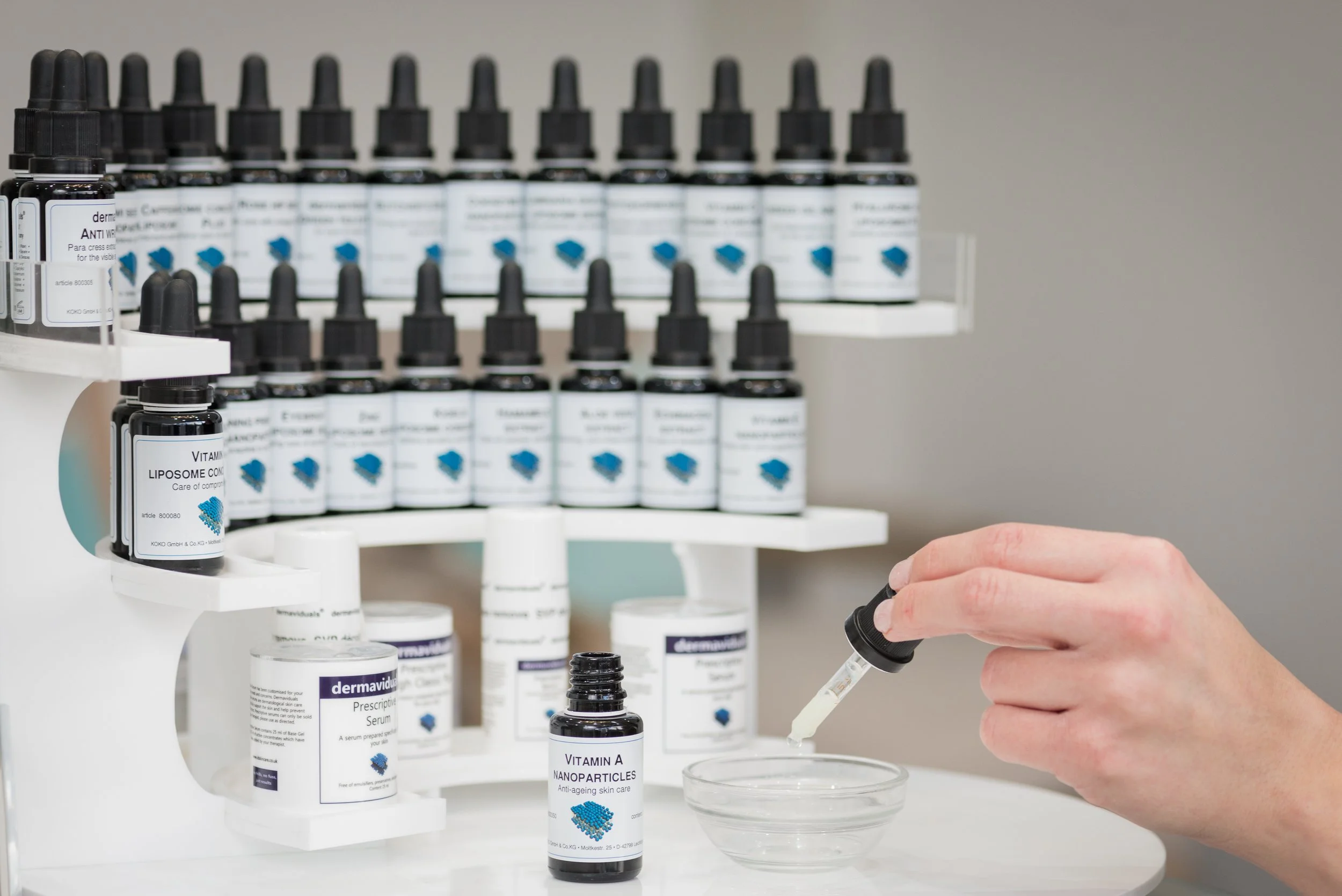 A person using a dropper to transfer liquid into a small clear bowl on a white table, with shelves of skincare bottles and containers in the background.