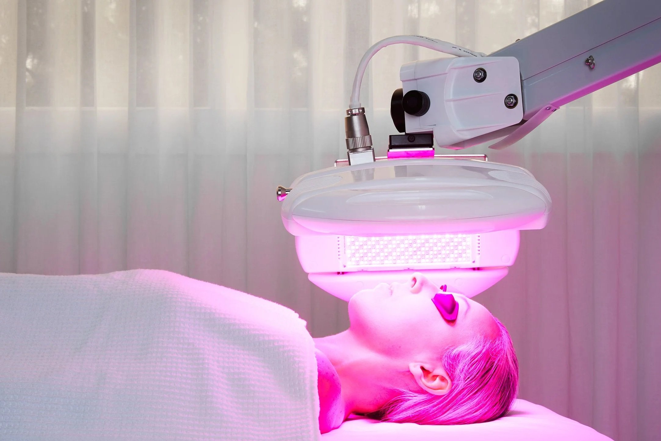 Lady lying down undergoing a medical or cosmetic procedure under a pink glow, with equipment positioned above the child's face.